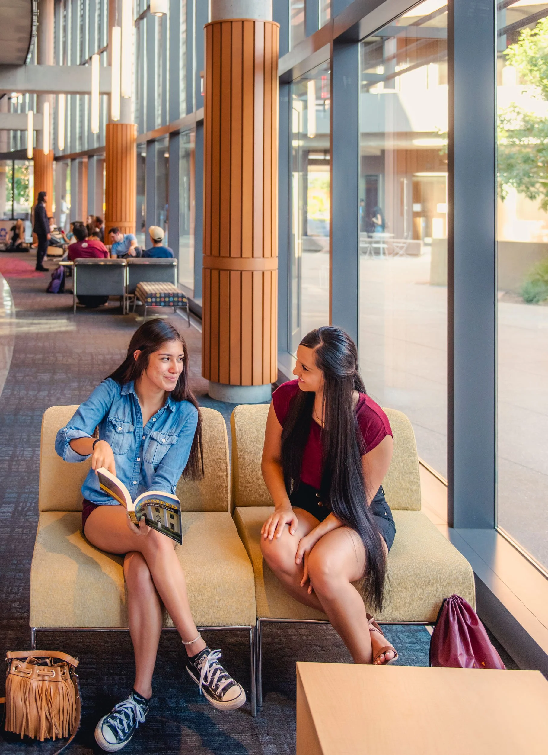 Two women sitting in a the Arizona State University library, talking. One is holding a book and the other has a smartphone on her lap in this Phoenix advertising photograph.