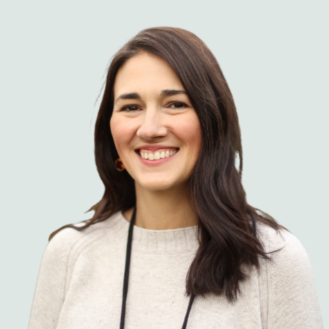 A woman with long dark brown hair smiling, wearing a light-colored sweater and a lanyard.