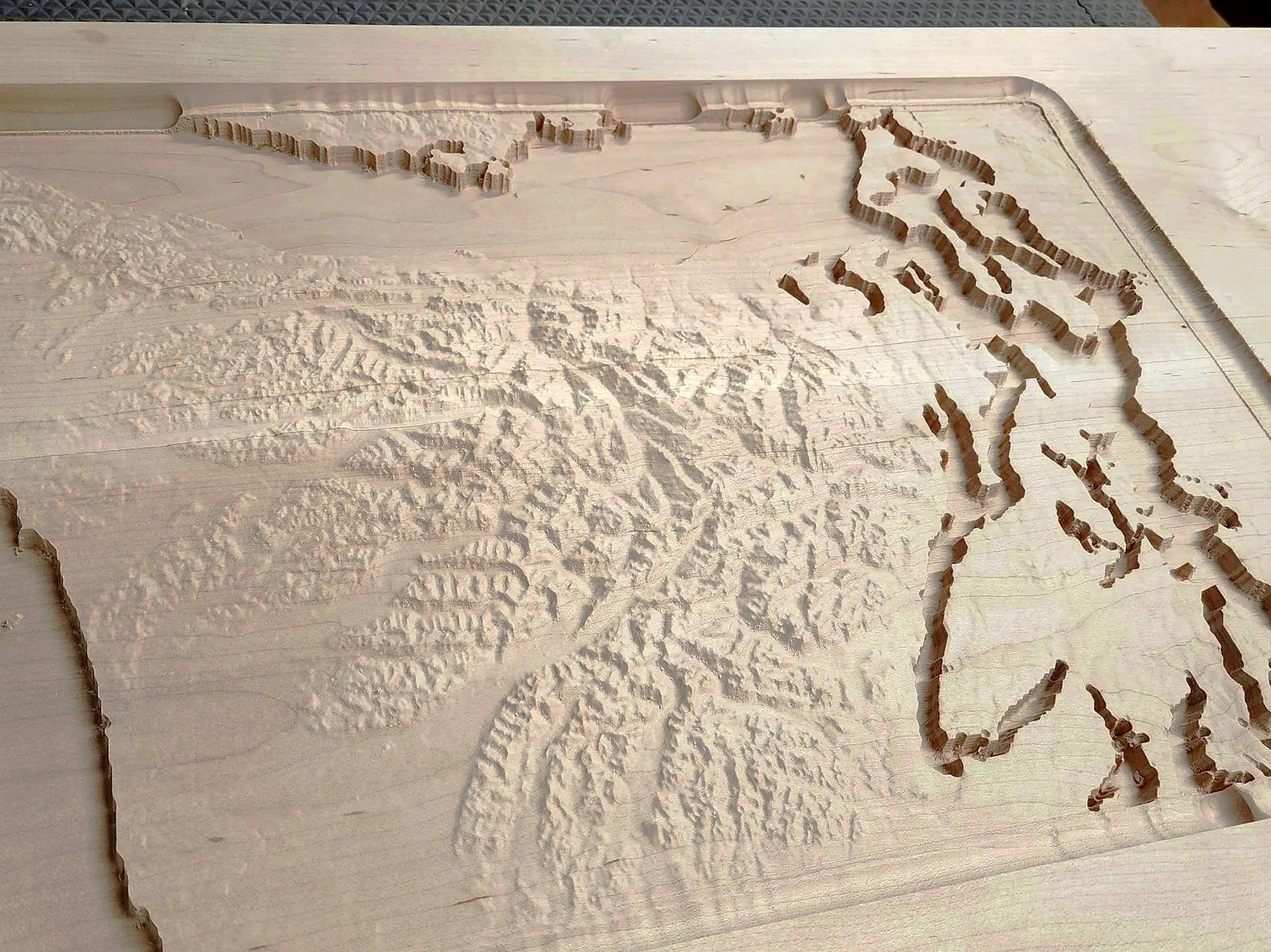 Wooden board with a carved topographical map, showing distinguishable landmasses and islands, surrounded by some equal-space indentations.