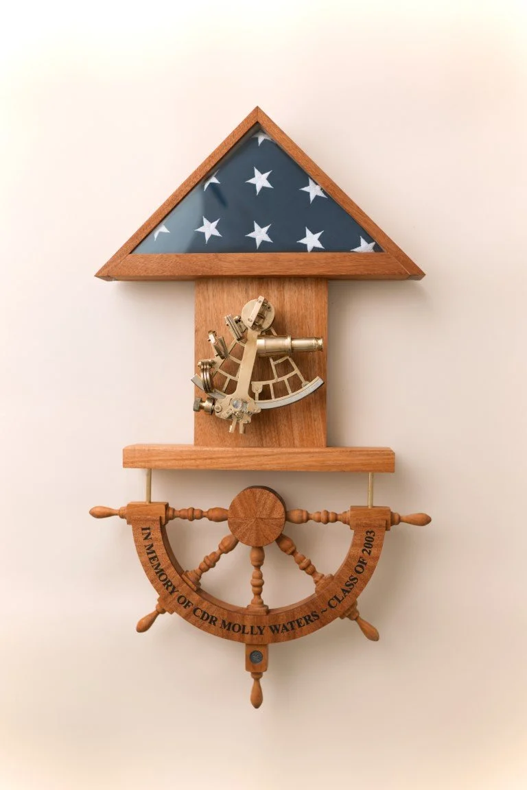 A wooden wall clock with a nautical theme, featuring a ship's wheel at the bottom, a telescope in the middle, and a triangular roof with a fabric patriotic pattern of stars and stripes.