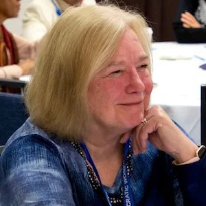 Professional woman with short black hair and glasses, wearing a white blazer, in an office setting.