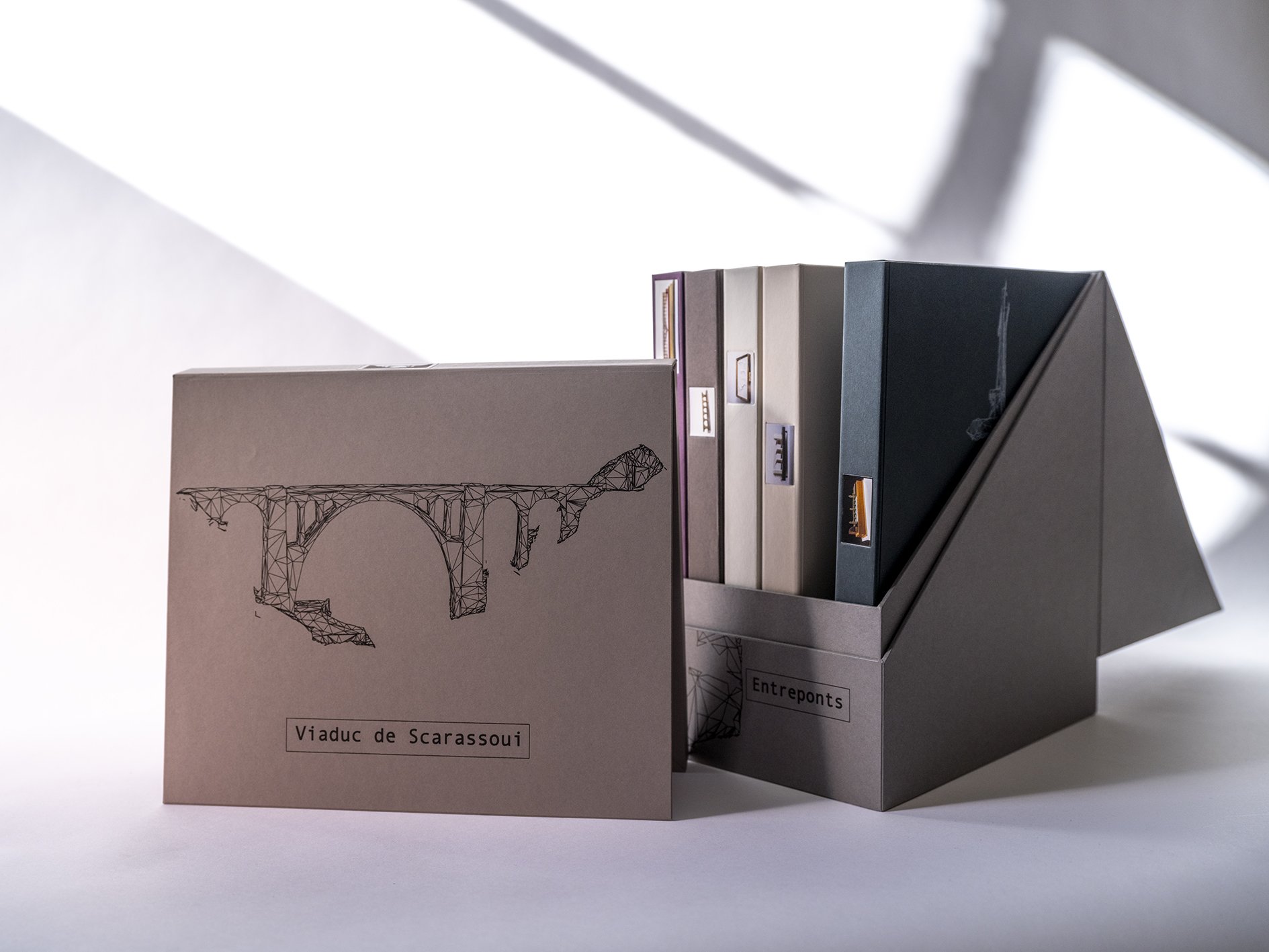 Coffret éditorial gris présentant une illustration de viaduc sur la couverture. À côté, un range-documents contient plusieurs livrets ou dossiers, évoquant un projet de recherche architecturale ou patrimoniale.