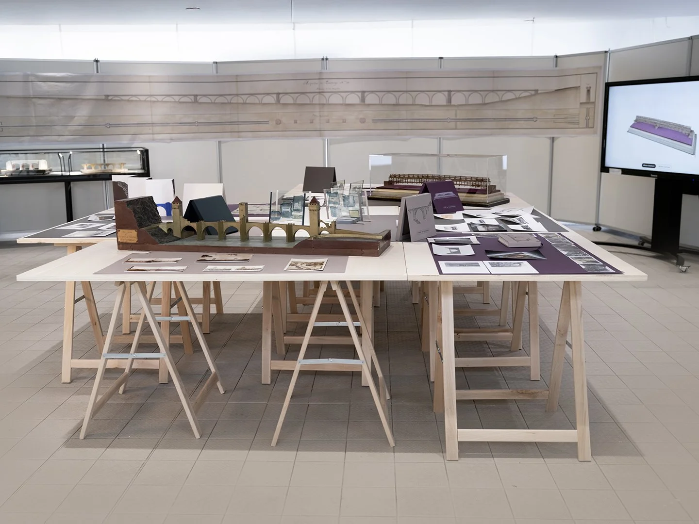 Espace d’exposition avec tables en bois présentant maquettes architecturales, plans et documents. Une maquette de pont à arches est au centre, avec en arrière-plan un long dessin technique mural et un écran d’analyse.