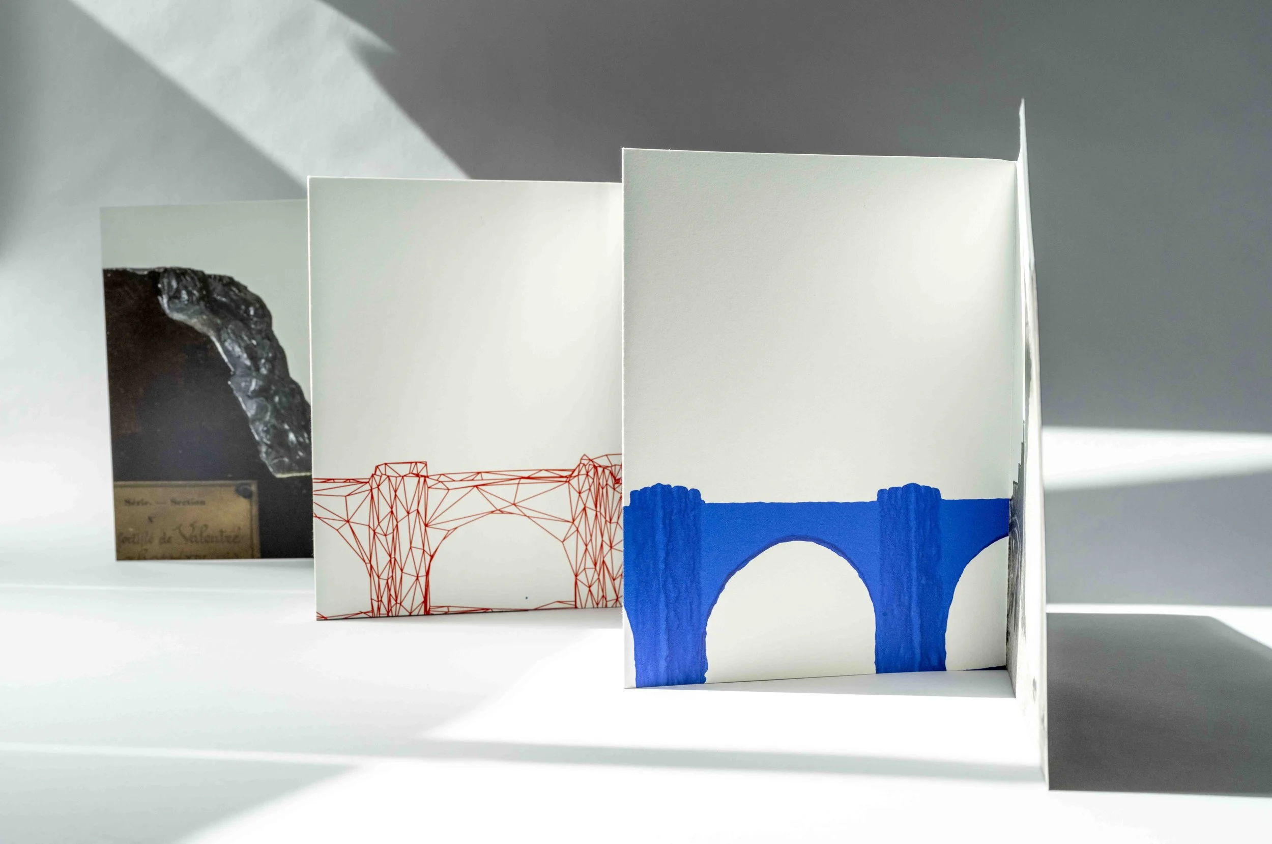 Édition imprimée composée de livrets ouverts aux couvertures blanches, ornées de dessins stylisés de ponts en rouge et bleu. L’ensemble est posé sur une surface claire, dans une mise en scène minimaliste.