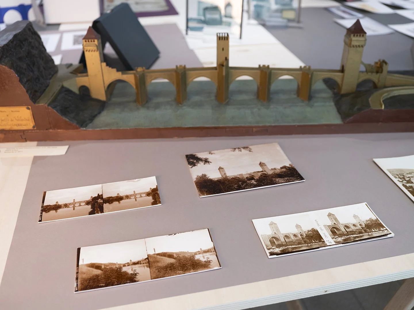 Maquette d’un pont fortifié à arches et tours, posée sur une table d’exposition. Devant, des photographies anciennes en sépia montrent le même pont dans son paysage. L’ensemble évoque une présentation historique et architecturale.