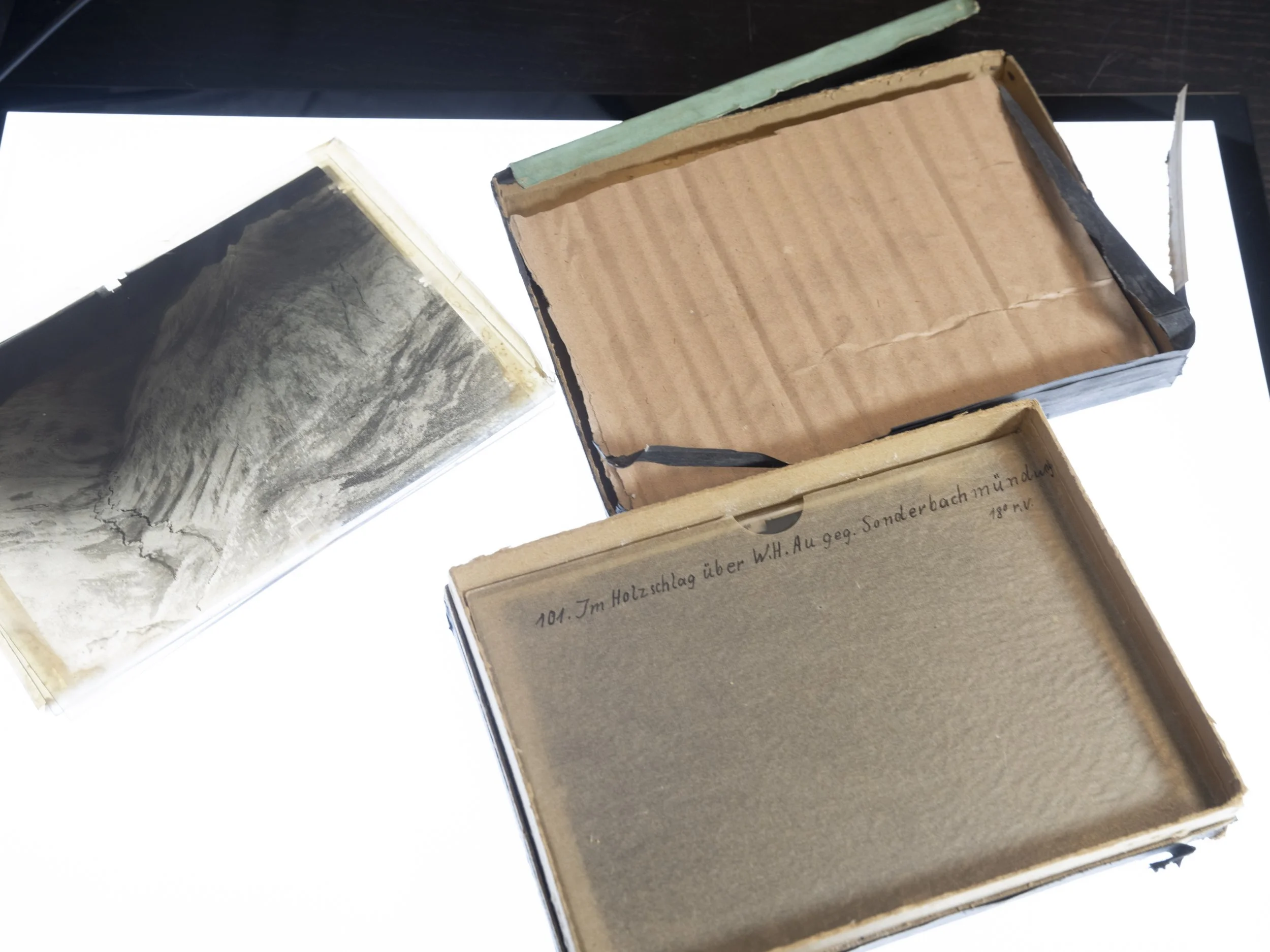 Trois boîtes d’archives anciennes ouvertes, en carton usé, posées sur une table lumineuse. L’une contient une photographie ancienne, les autres semblent vides ou portent des annotations manuscrites.