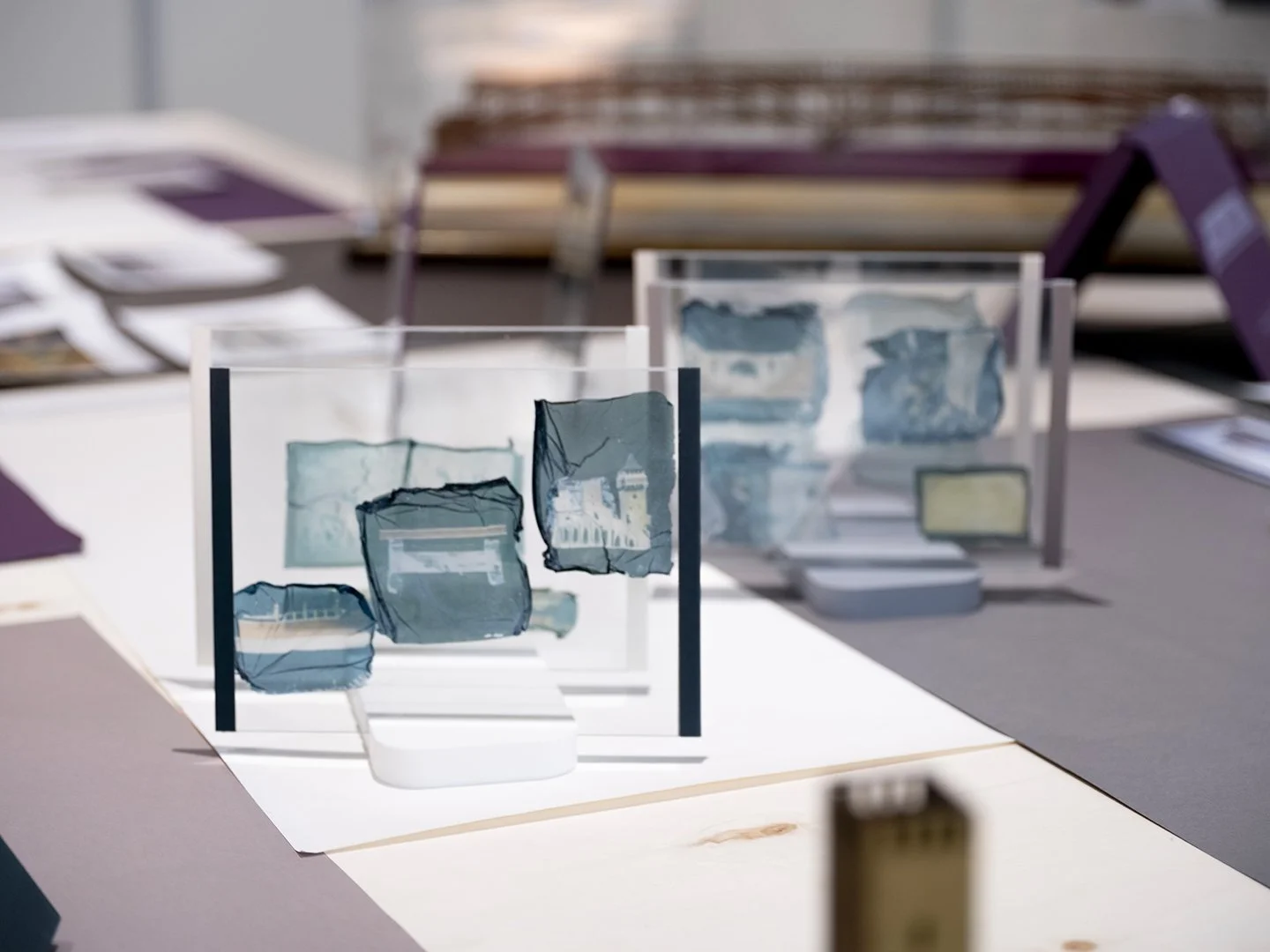 Espace d’exposition avec tables en bois présentant maquettes architecturales, plans et documents. Une maquette de pont à arches est au centre, avec en arrière-plan un long dessin technique mural et un écran d’analyse.