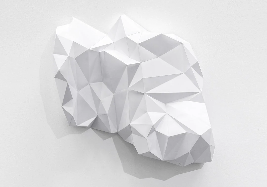 Sculpture murale abstraite blanche composée de facettes géométriques anguleuses. Le volume en relief crée des jeux d’ombres subtils sur un mur blanc, évoquant une forme minérale ou topographique dans un style minimaliste.