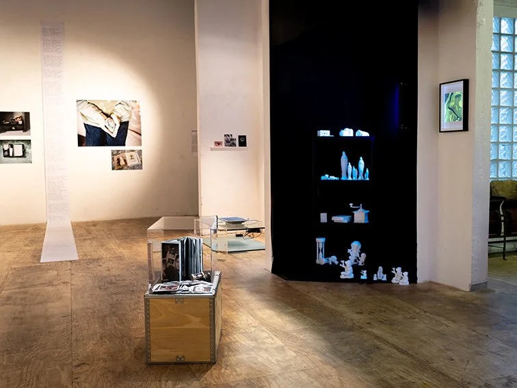 Espace d’exposition contemporain avec parquet en bois, photos accrochées aux murs et objets protégés sous vitrines. Une niche sombre illuminée révèle des sculptures blanches aux formes variées.