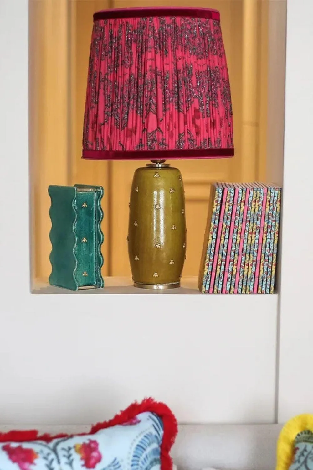 une lampe à poser montrée dans une niche, avec un pied en céramique couleur moutarde et un abat jour rose