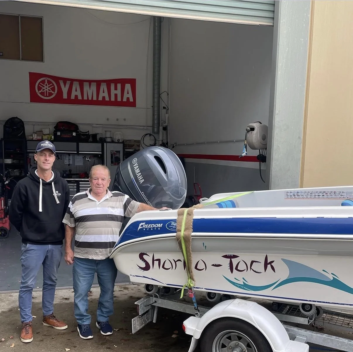 Two men standing next to a small boat with a Yamaha outboard motor in a garage with Yamaha branding.