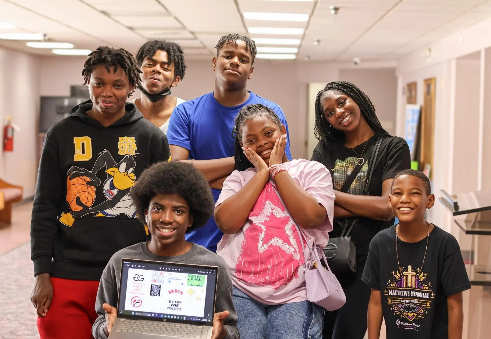 Group of nine diverse young people gathered indoors, some smiling and some making expressive faces, with one person holding a laptop showing various graphics and text.
