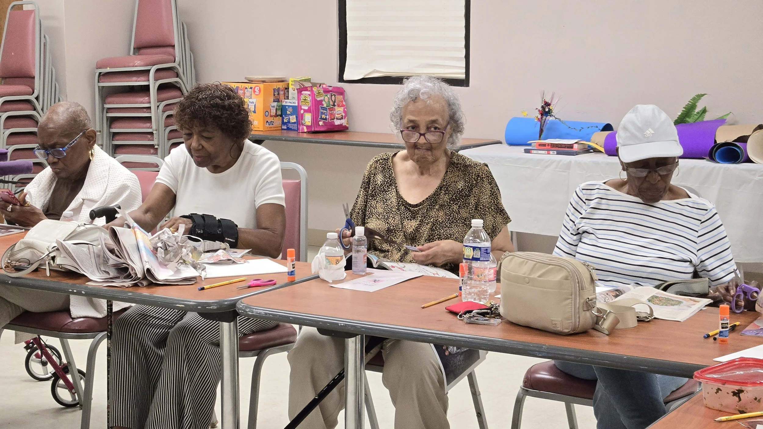 Four elderly women sitting at a table engaged in arts and crafts, with supplies such as scissors, glue, papers, and notebooks, in a community room.