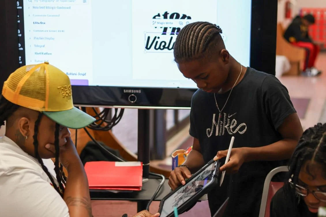 Two young people working on a tablet in front of a large screen displaying a font design project, with another young person visible in the background.
