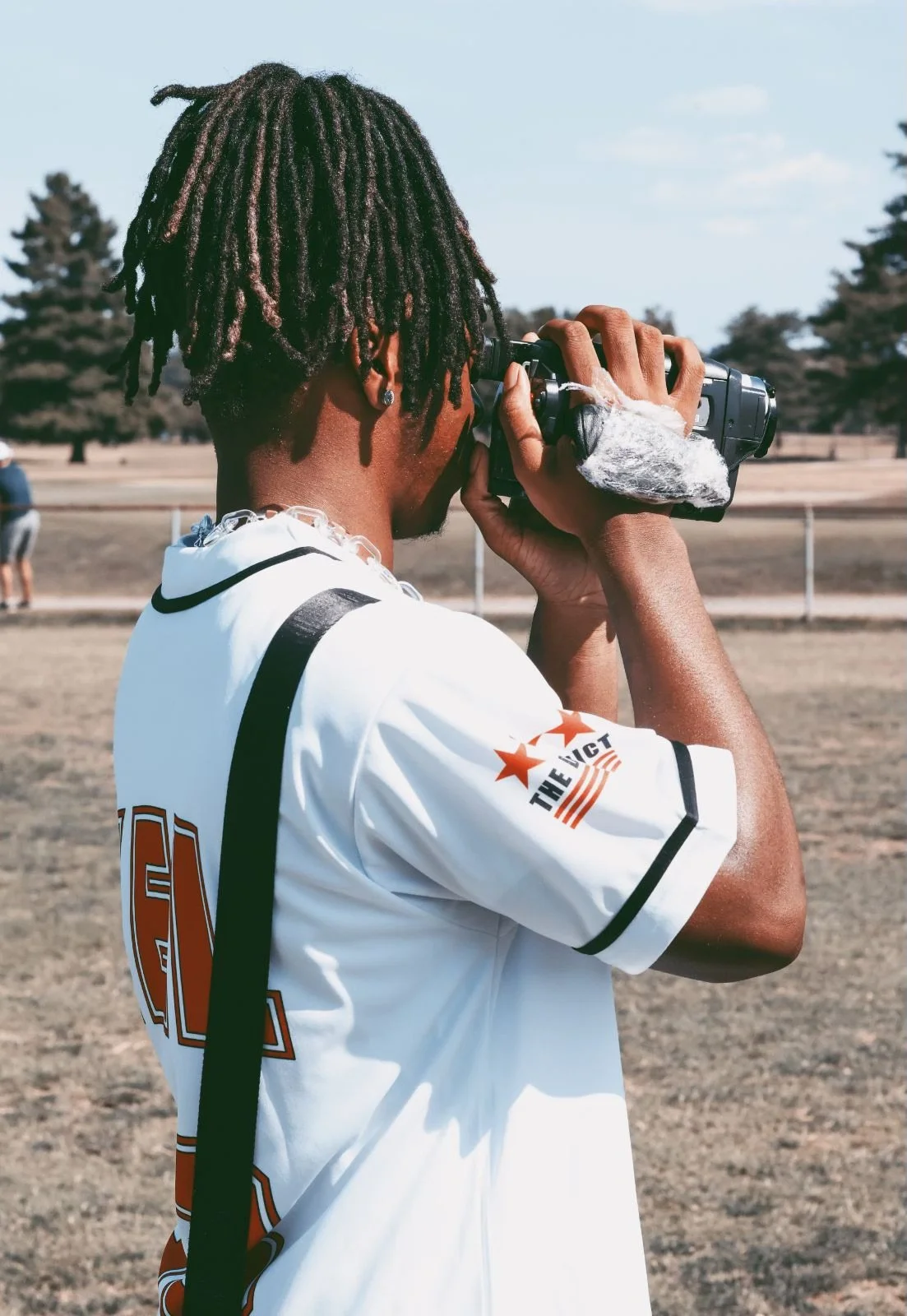 A young man with dreadlocks wearing a white sports jersey taking a photograph with a camera outdoors in a park with trees in the background.