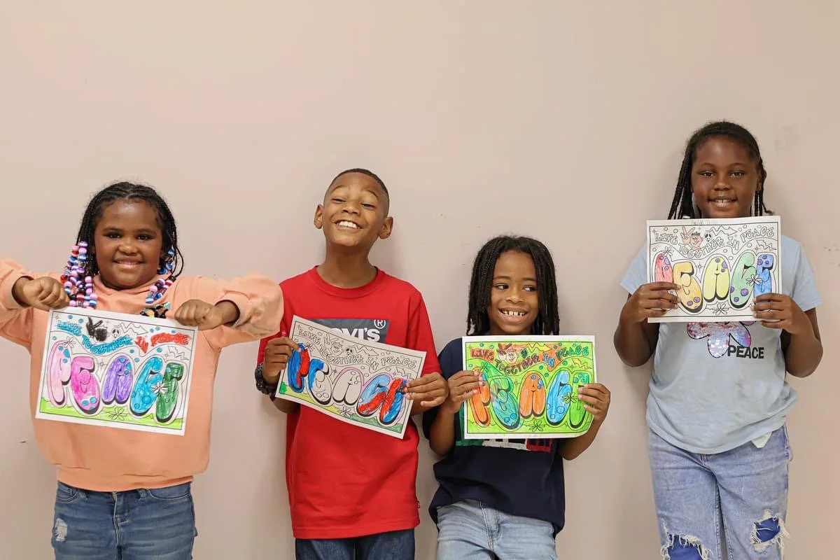 Four children standing against a plain wall, each holding a sign with the word 'peace' written in colorful, artistic letters, celebrating unity and peace.