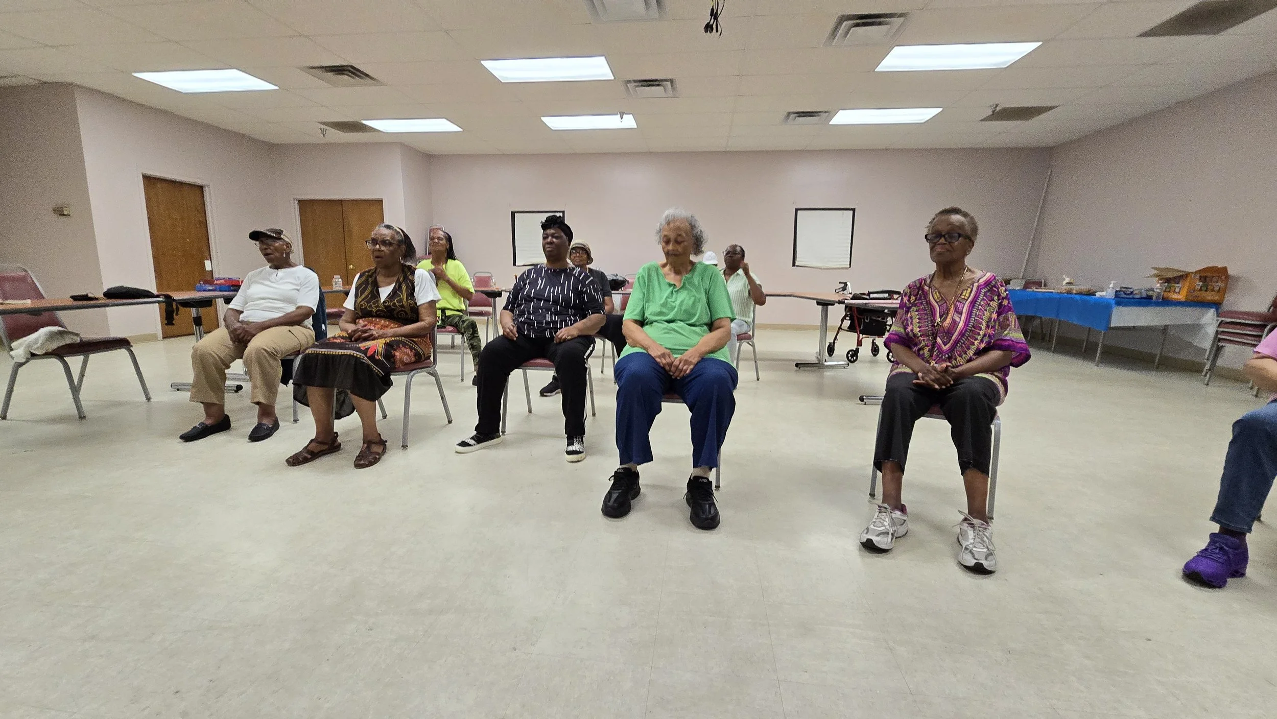 Group of elderly people sitting on chairs in a spacious room, likely attending a class or event, with tables and supplies in the background.