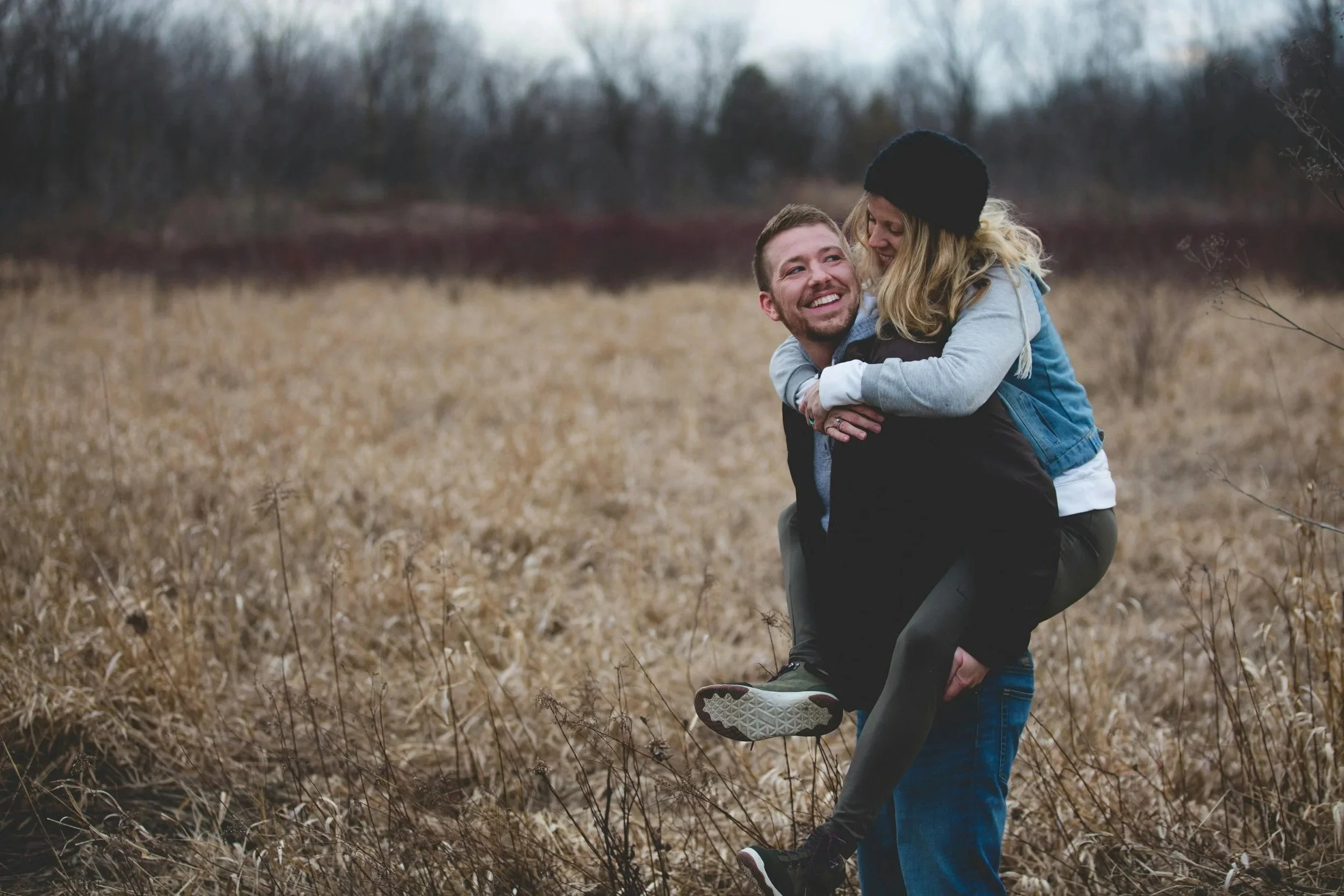 A couple outdoors, woman on man's back in a piggyback ride, smiling, in a field with dry grass and trees in the background.