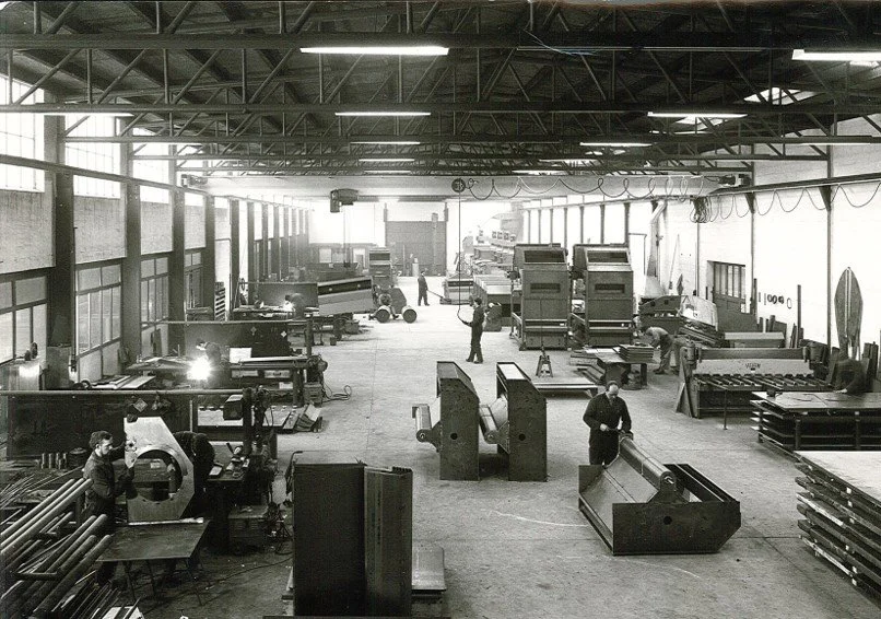 Черно-белая фотография большого промышленного цеха или завода, на которой рабочие обслуживают станки и собирают металлические детали.