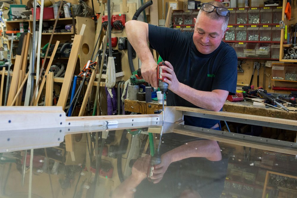 A man in a workshop, assembling or securing a piece of wood using a power drill. The workspace is filled with tools, wood pieces, and shelves.