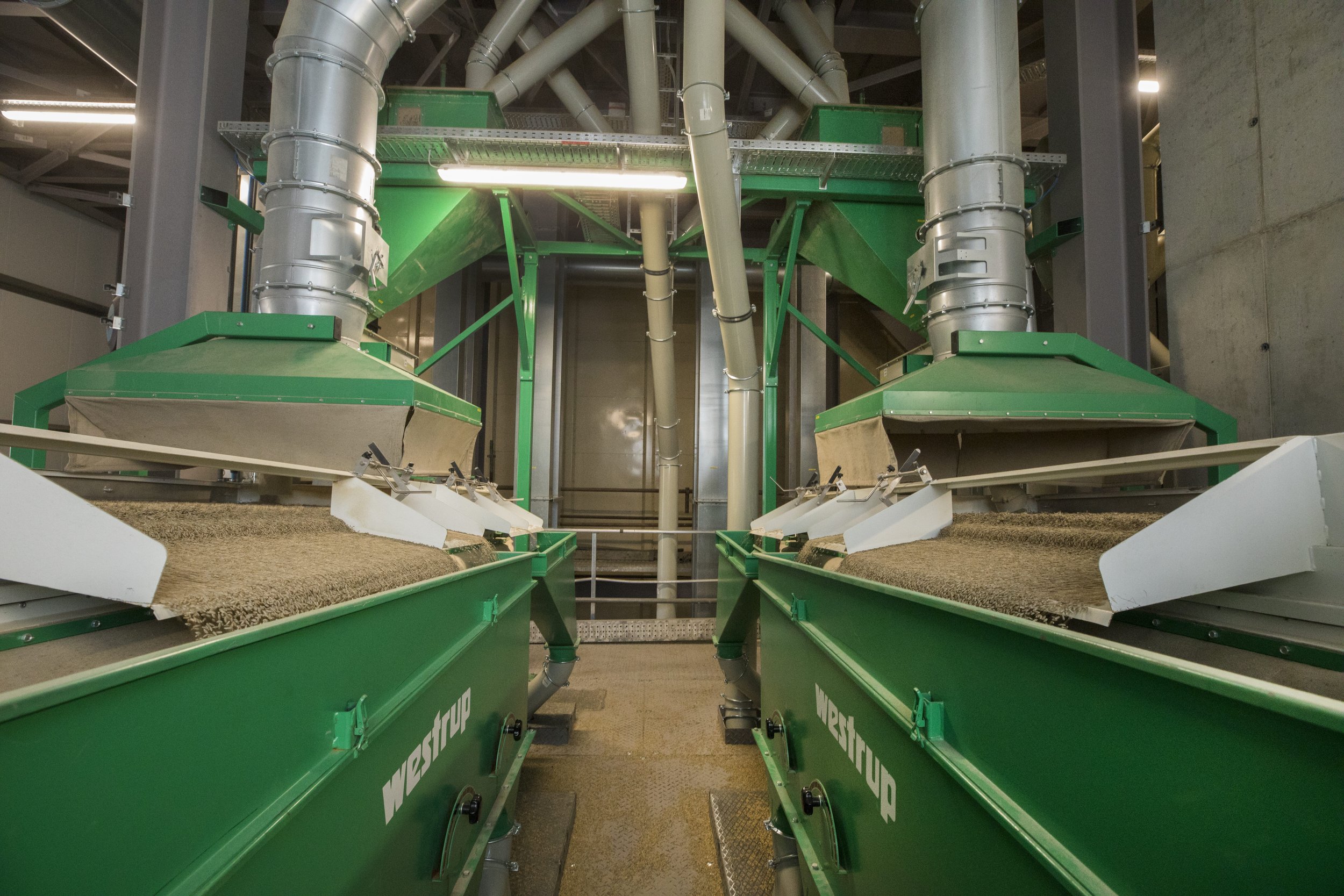Industrial machinery used for processing grains or seeds at a large facility. The equipment features green metal chutes and conveyors with beige burlap-like coverings, surrounded by large pipes and metal infrastructure.