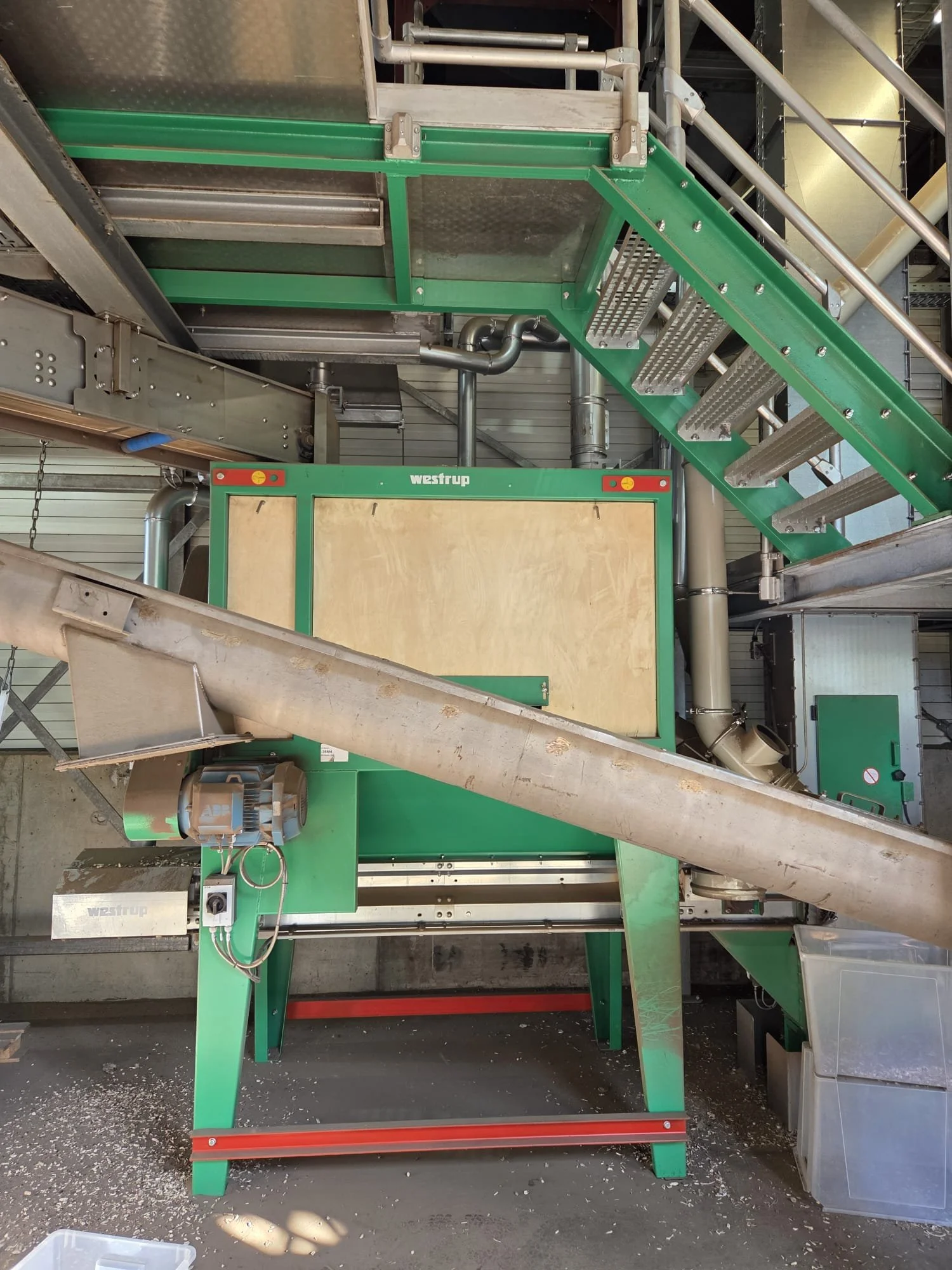 Industrial machinery including a green conveyor system, metal stairs, pipes, and a large wooden panel in a warehouse setting.