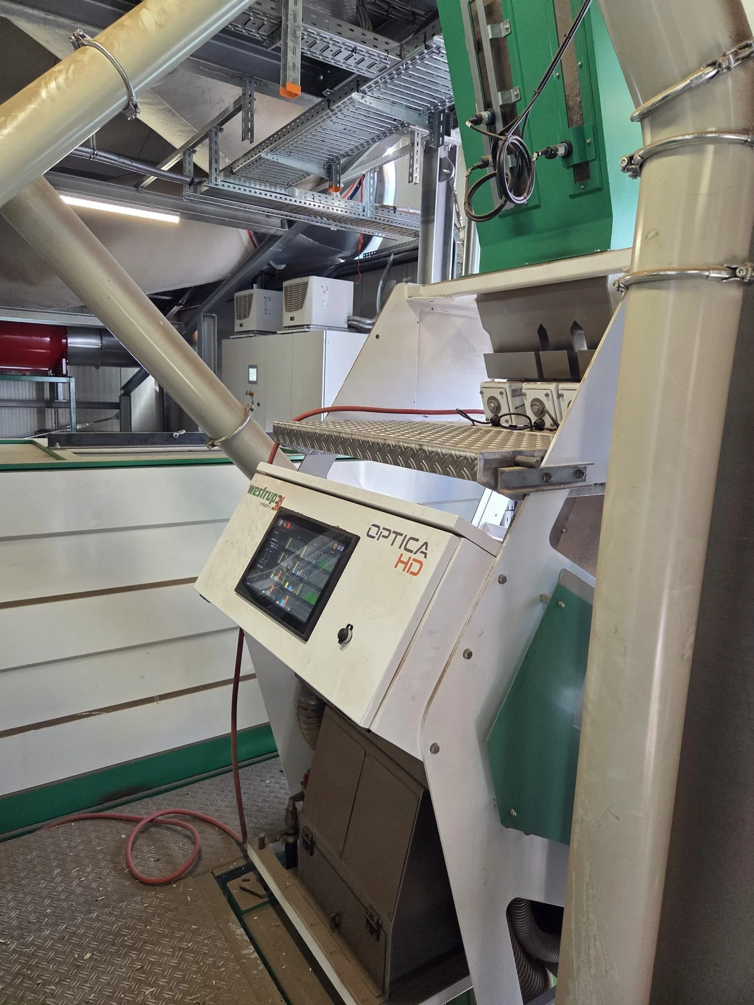 Close-up of an industrial machine with a touchscreen interface labeled 'Optica HD,' in a manufacturing or processing plant. The machine has a white exterior with green accents and is surrounded by metal pipes, frames, and electrical components.