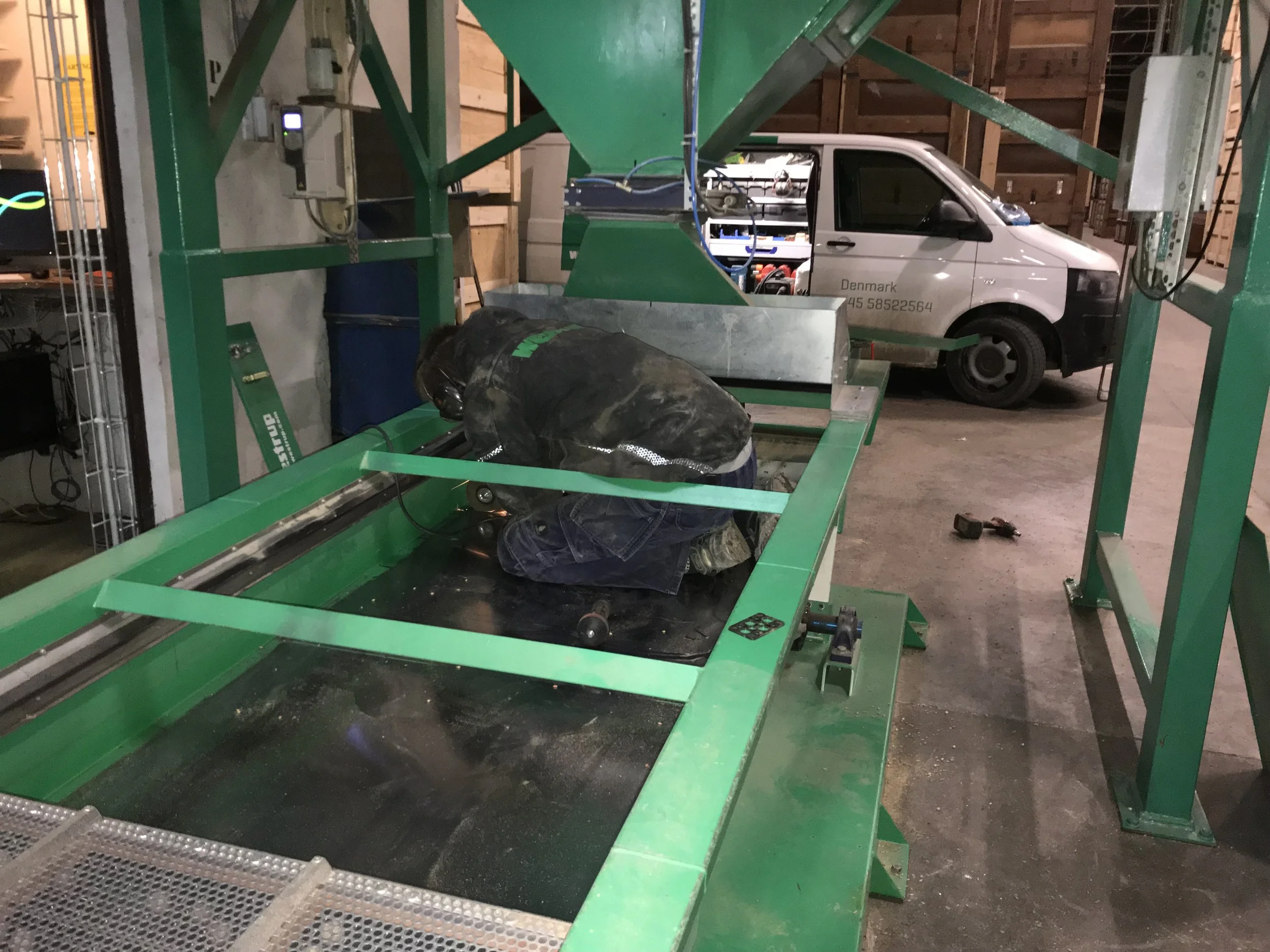 Eine Person, die in einem Lager oder einer Fabrik an Maschinen arbeitet. Die Person kniet und scheint an der grünen Maschinen zu arbeiten oder diese zu reparieren. Im Hintergrund ist ein kleiner weißer Lieferwagen zu sehen, und der Bereich ist von Holzwänden umgeben.