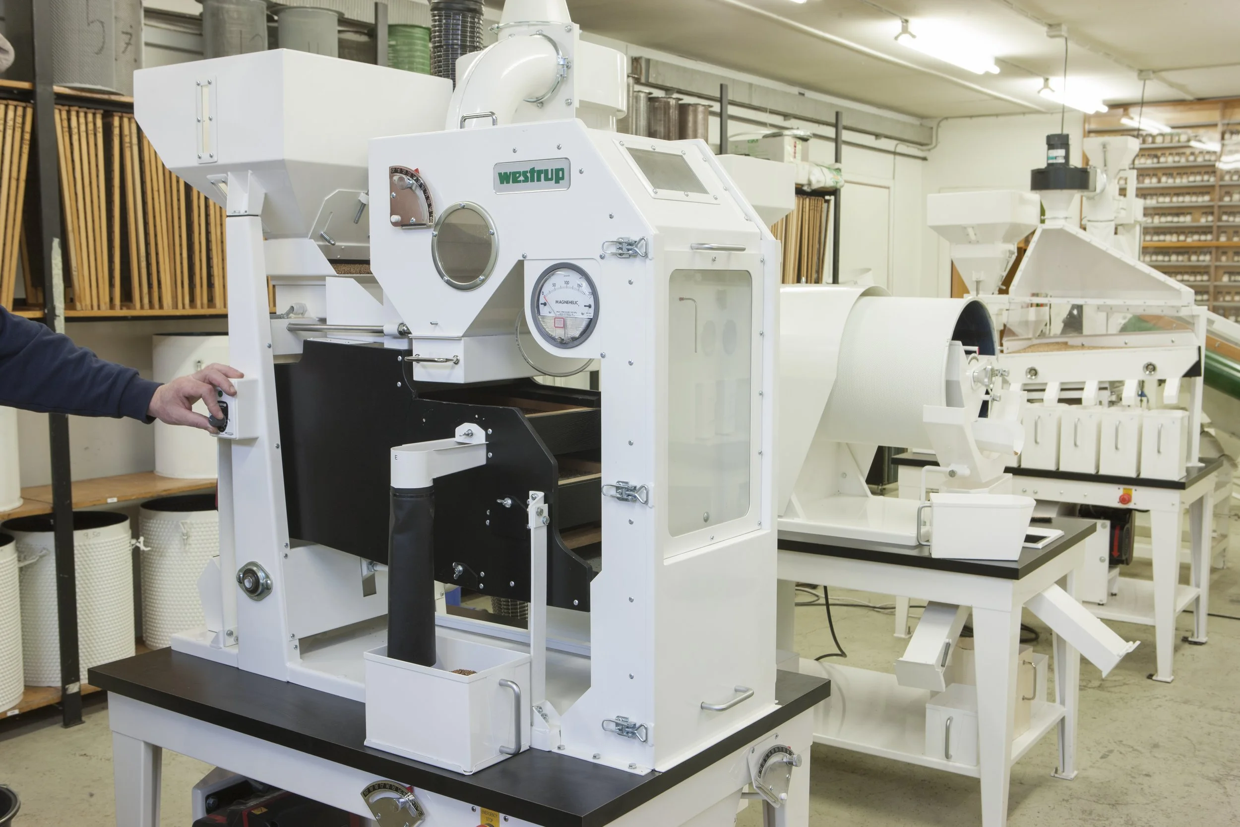 Промышленная машина, используемая для производства или переработки на заводе; на левой стороне человек вручную регулирует органы управления, вокруг — полки и другое оборудование.