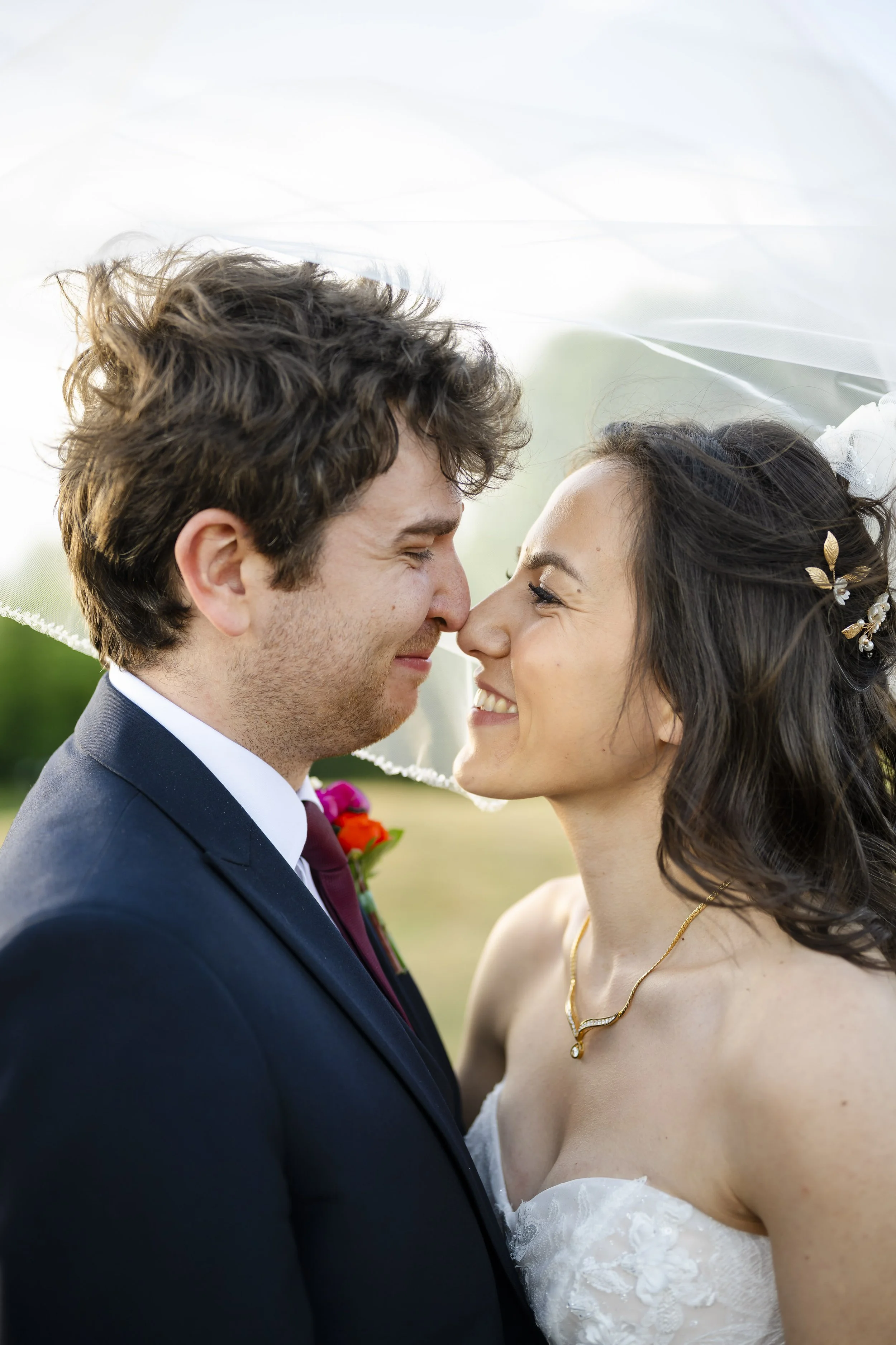 A newlywed couple touching noses outdoors, smiling, with the bride wearing a veil, white lace dress, and gold necklace, and the groom in a dark suit and maroon tie, in a romantic moment.