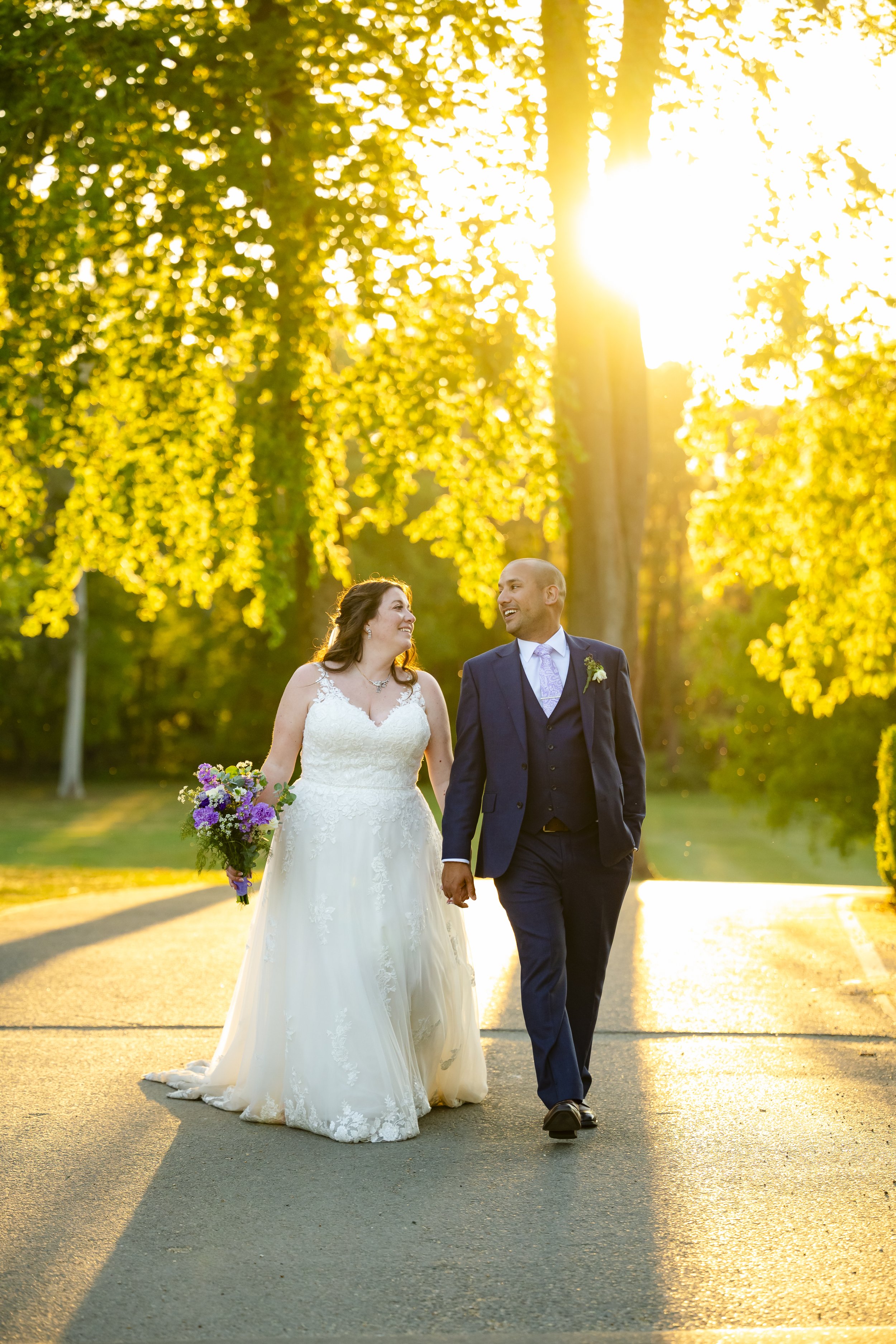 A newlywed couple walking hand in hand on a tree-lined street during sunset, with the bride in a wedding gown holding a bouquet, and the groom in a dark suit.