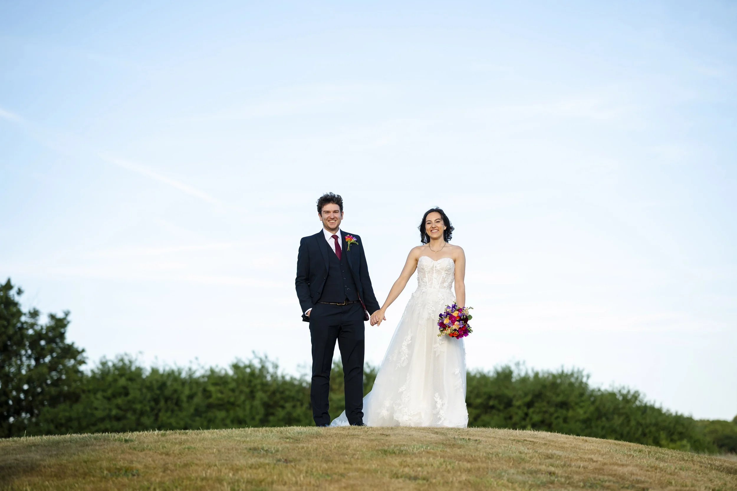 A bride and groom holding hands, standing on a grassy hill outdoors with a clear sky in the background.