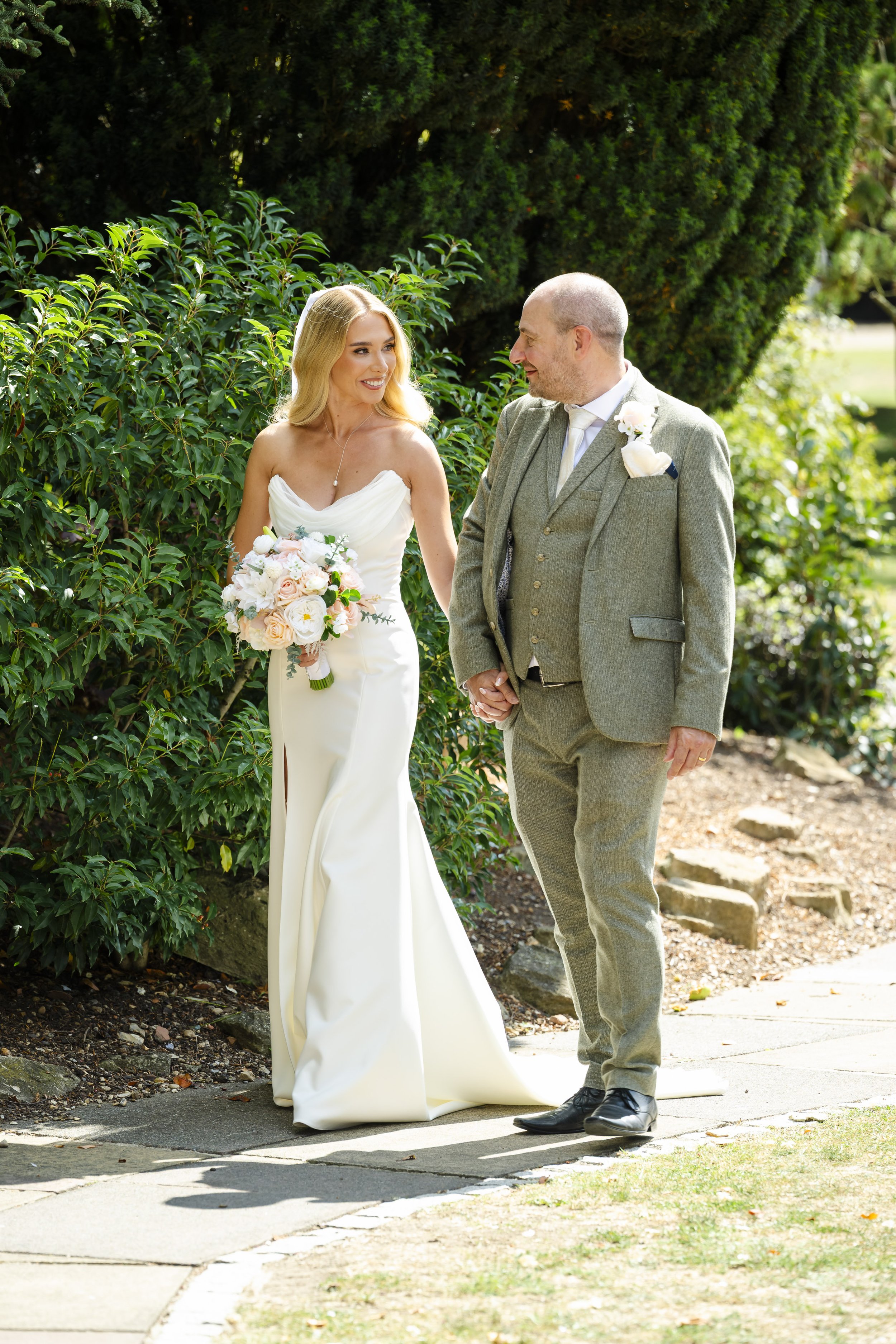 A bride and an older man, likely her father, walking outdoors in a garden, holding hands and smiling at each other, with greenery and bushes in the background.