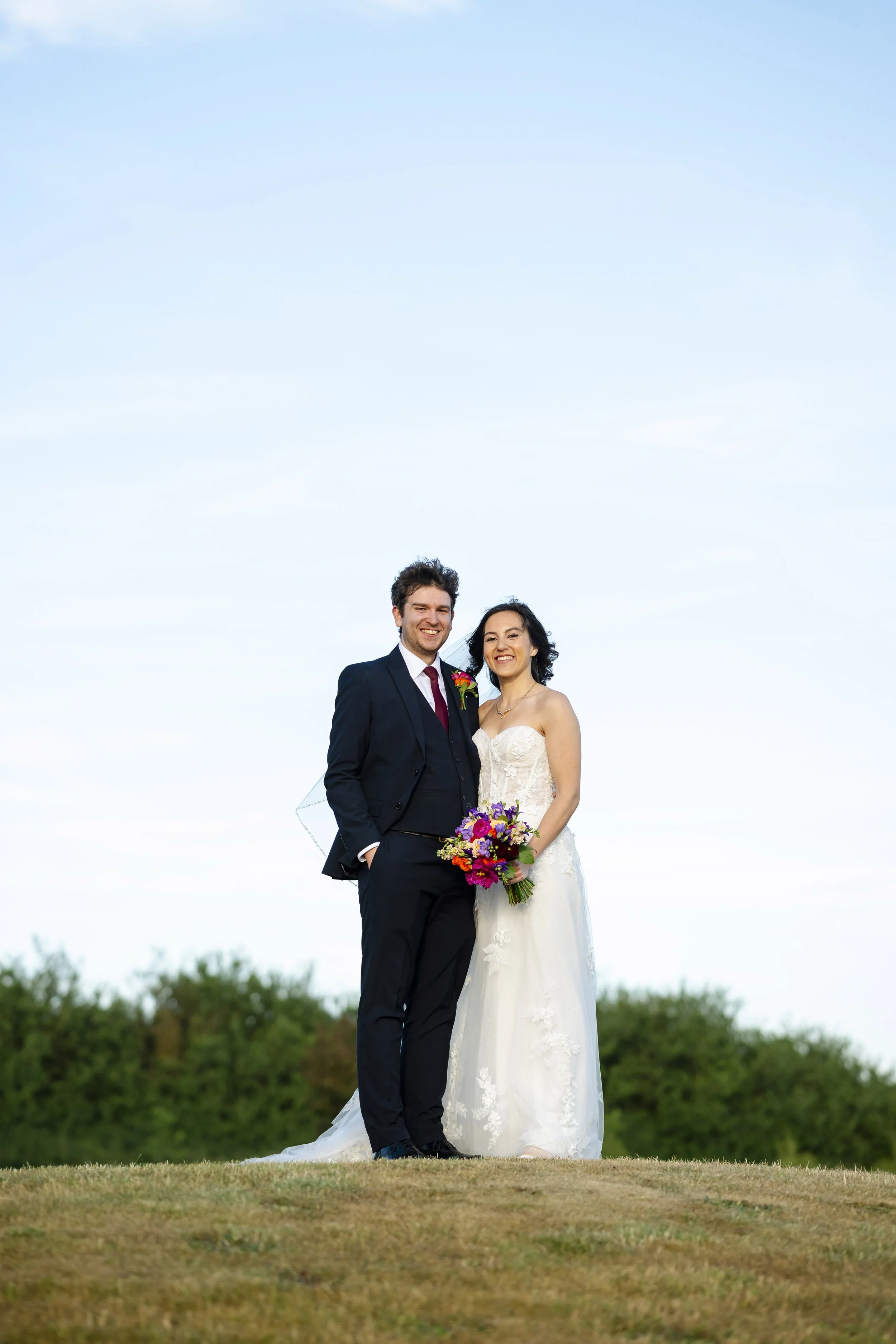 A newlywed couple in wedding attire standing outdoors on a grassy area, smiling at the camera with a clear sky and green trees in the background.