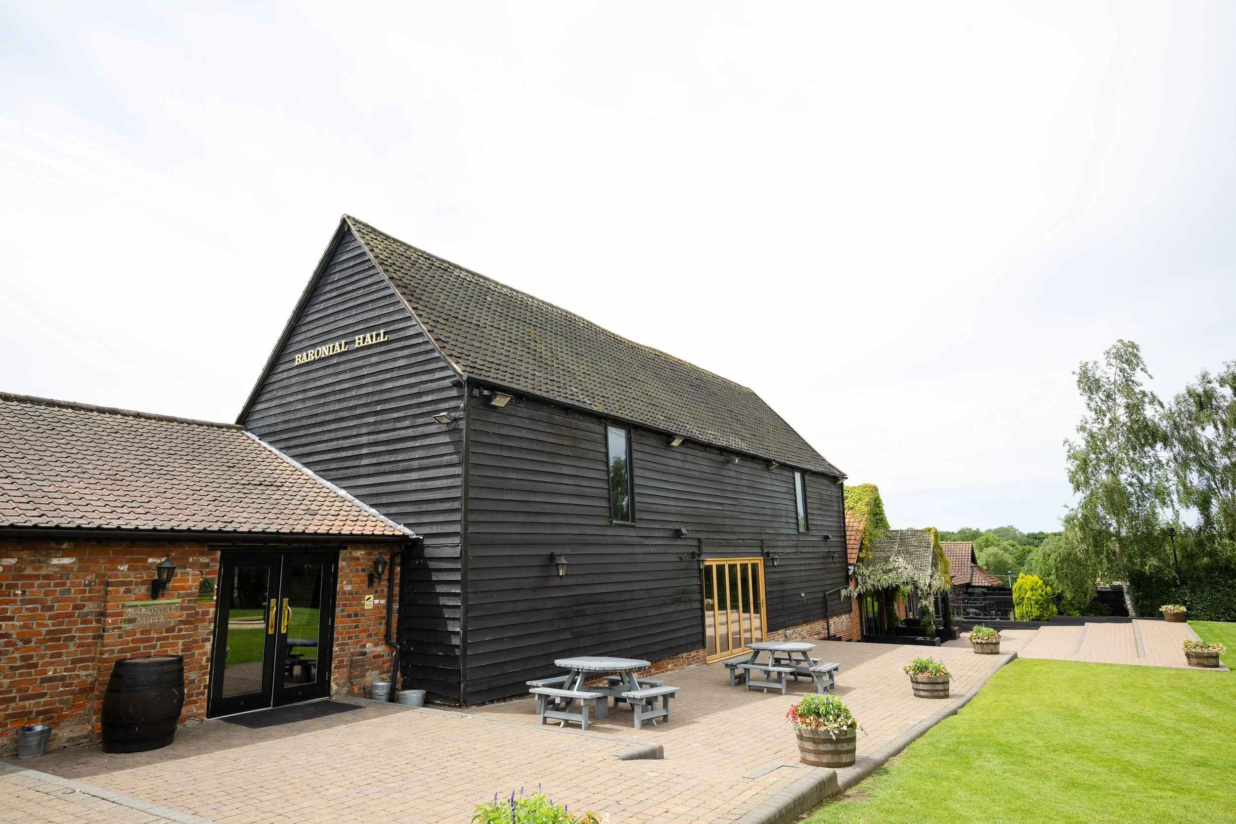 Exterior of Baronal Hall, a building with black wooden siding and a brick section, surrounded by a paved patio with picnic tables and decorative planters, and a green lawn with trees in the background.