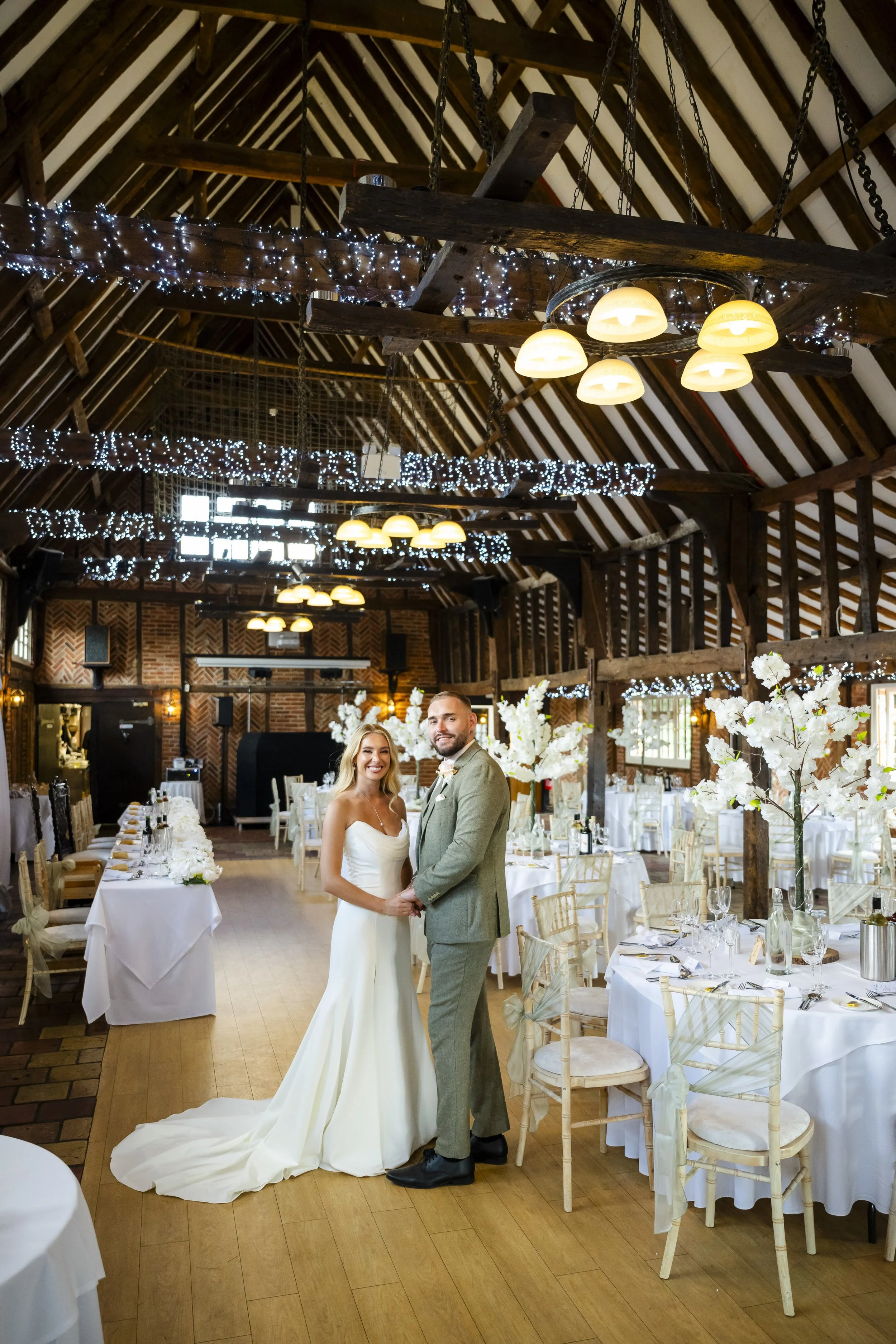 A bride and groom stand hand in hand in a decorated wedding reception hall with wooden beams, string lights, and floral centerpieces.