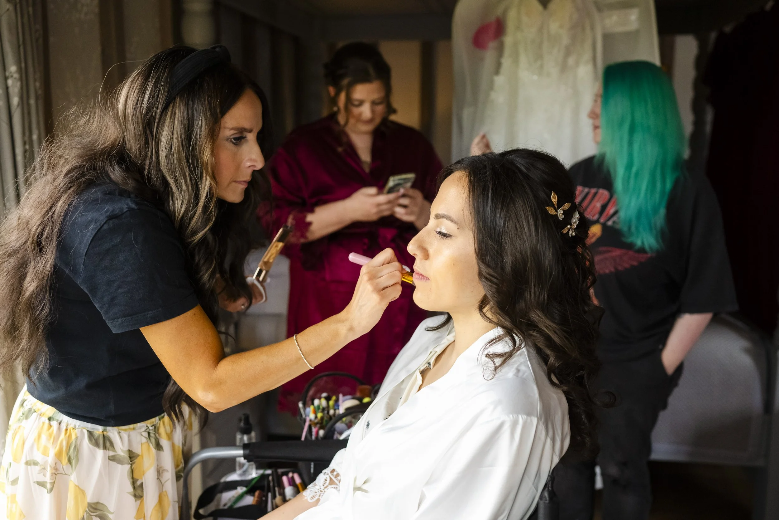 A woman with long dark hair sitting with eyes closed while a makeup artist applies lipstick on her lips. Other women are in the background, one with green hair and another using a phone, in a room with a wedding dress hanging nearby.