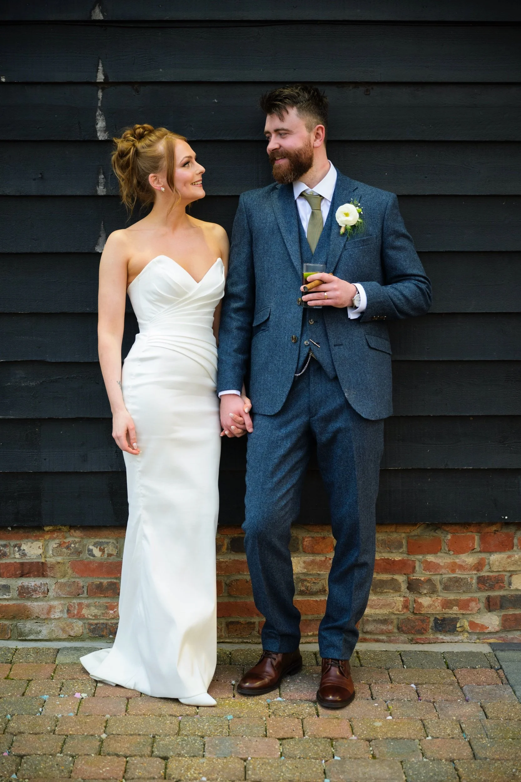 A bride and groom holding hands, looking at each other, standing against a black wooden wall. The bride is in a strapless white wedding gown, and the groom is in a blue suit holding a drink.