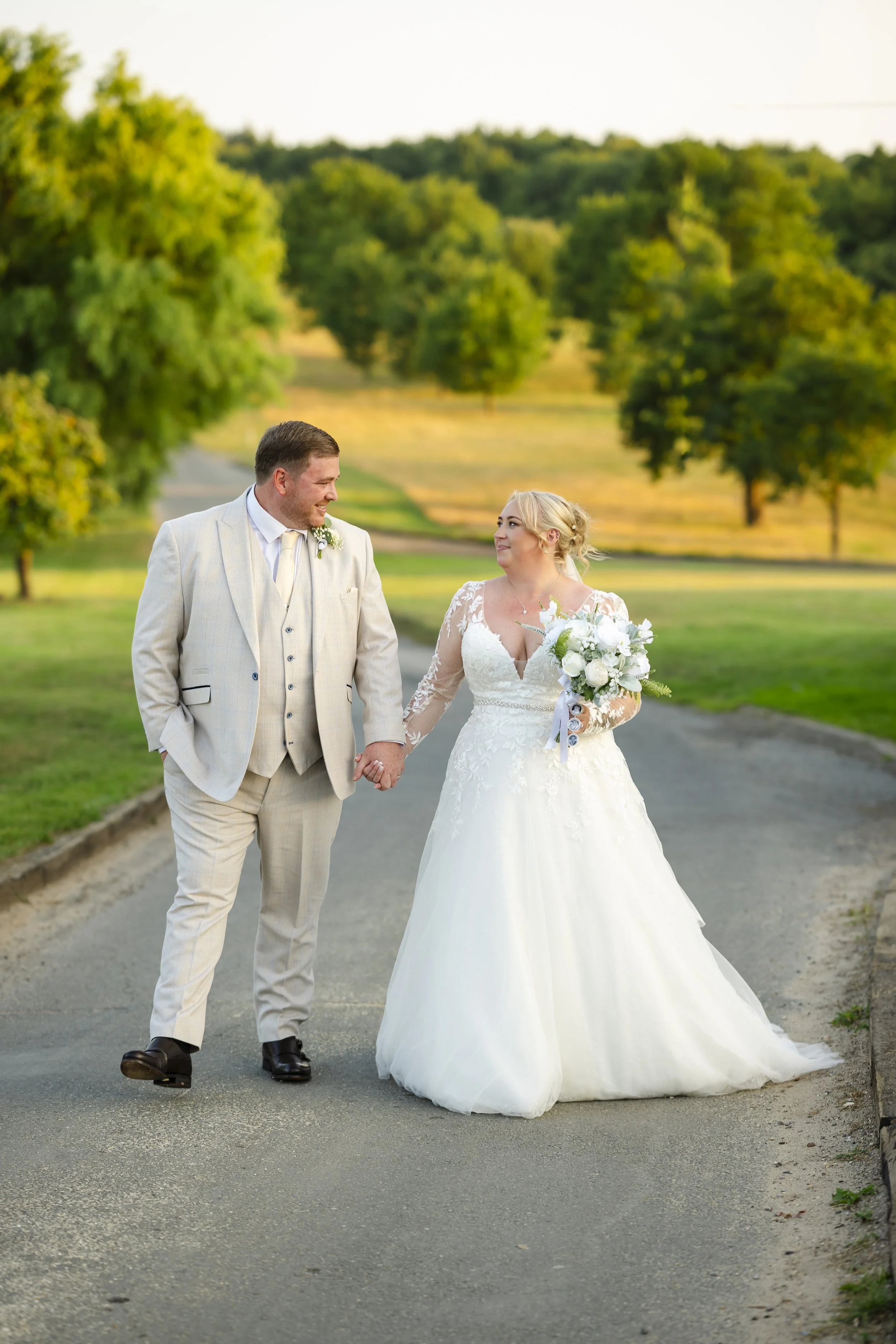A bride and groom walking hand in hand outdoors during sunset on a paved path, with trees and grassy fields in the background.