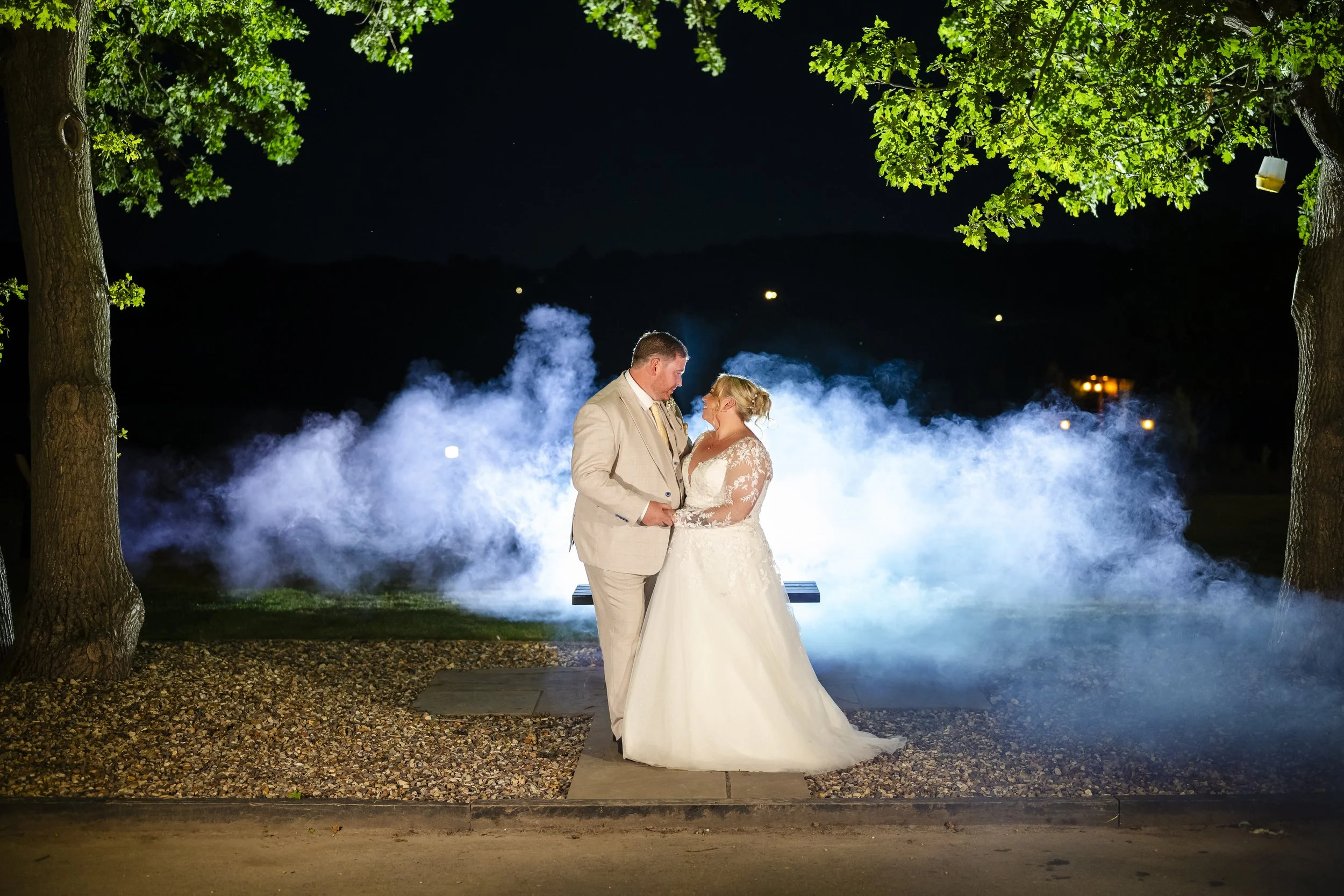 A bride and groom in wedding attire standing together at night outdoors, with fog and bright lights in the background, framed by large trees.