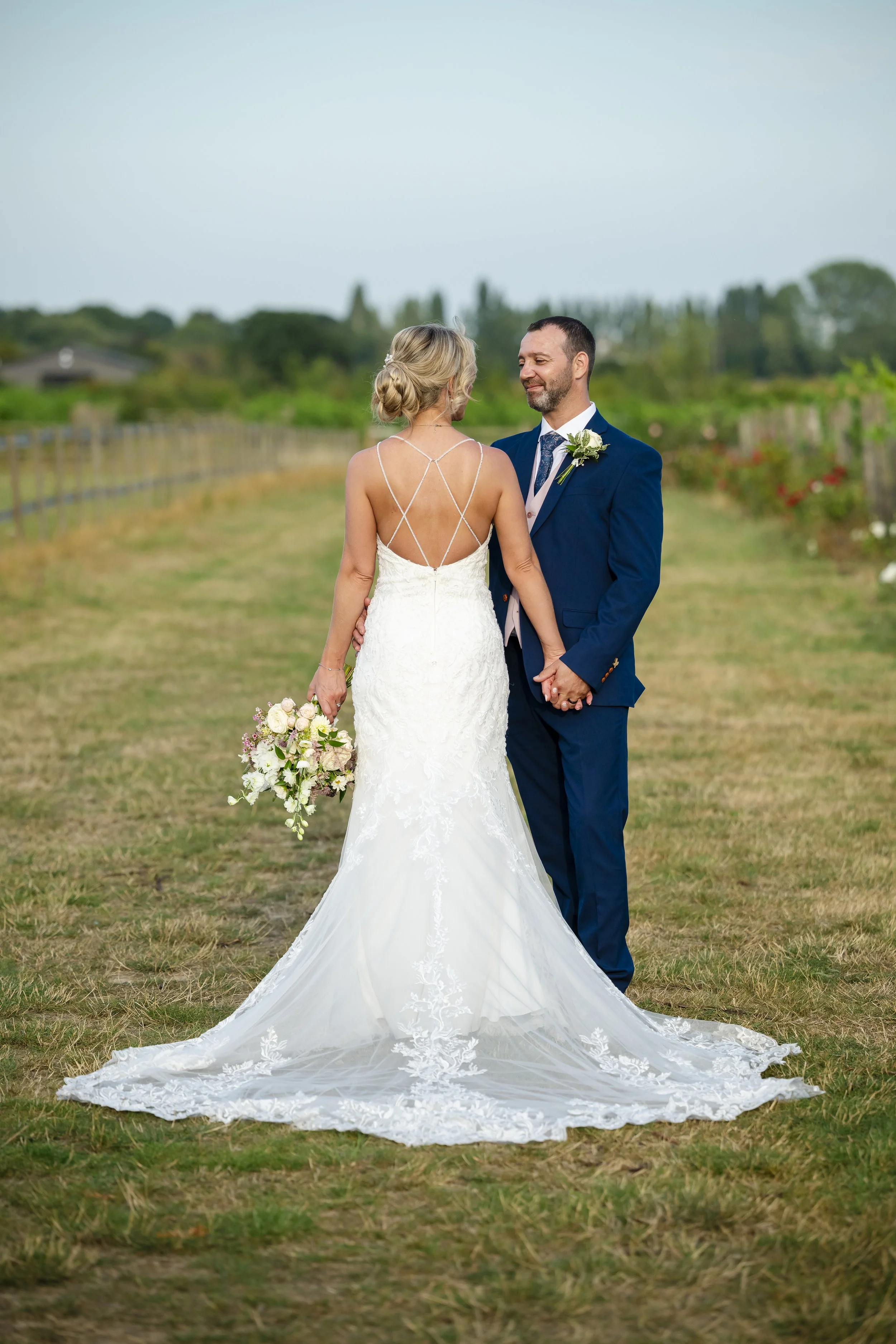 A bride and groom holding hands outdoors on their wedding day, facing each other with a garden and cloudy sky in the background.