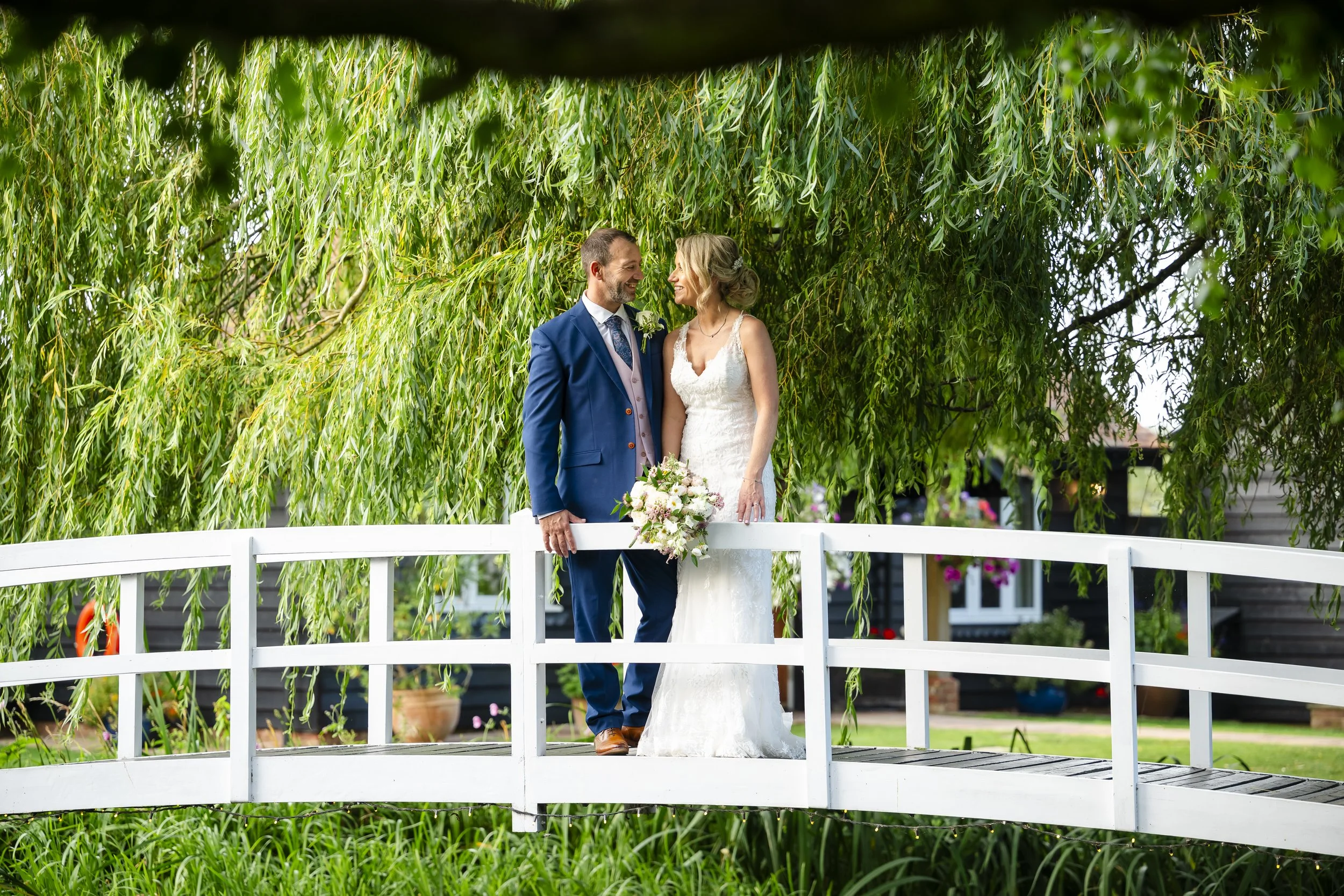 A bride and groom standing on a white bridge surrounded by lush greenery, smiling at each other with the bride holding a bouquet of flowers.