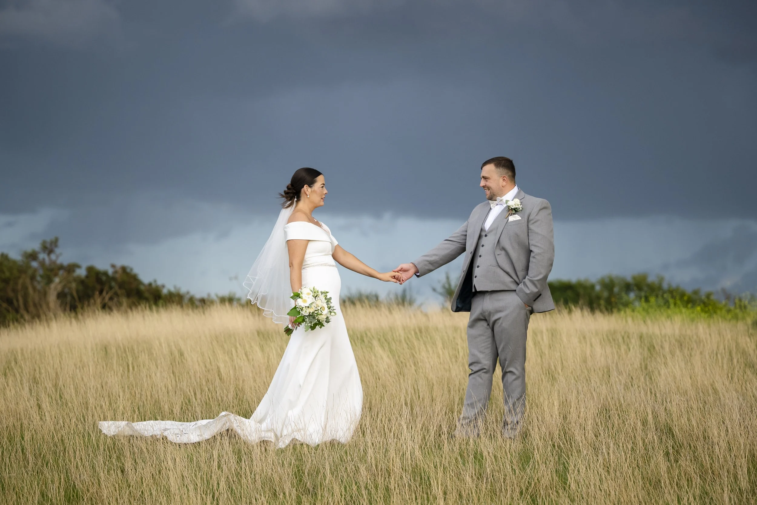 A bride and groom standing in a grassy field holding hands. The bride is in a white wedding gown with a veil, holding a bouquet of flowers. The groom is in a gray suit with a boutonnière. Dark storm clouds are in the background.