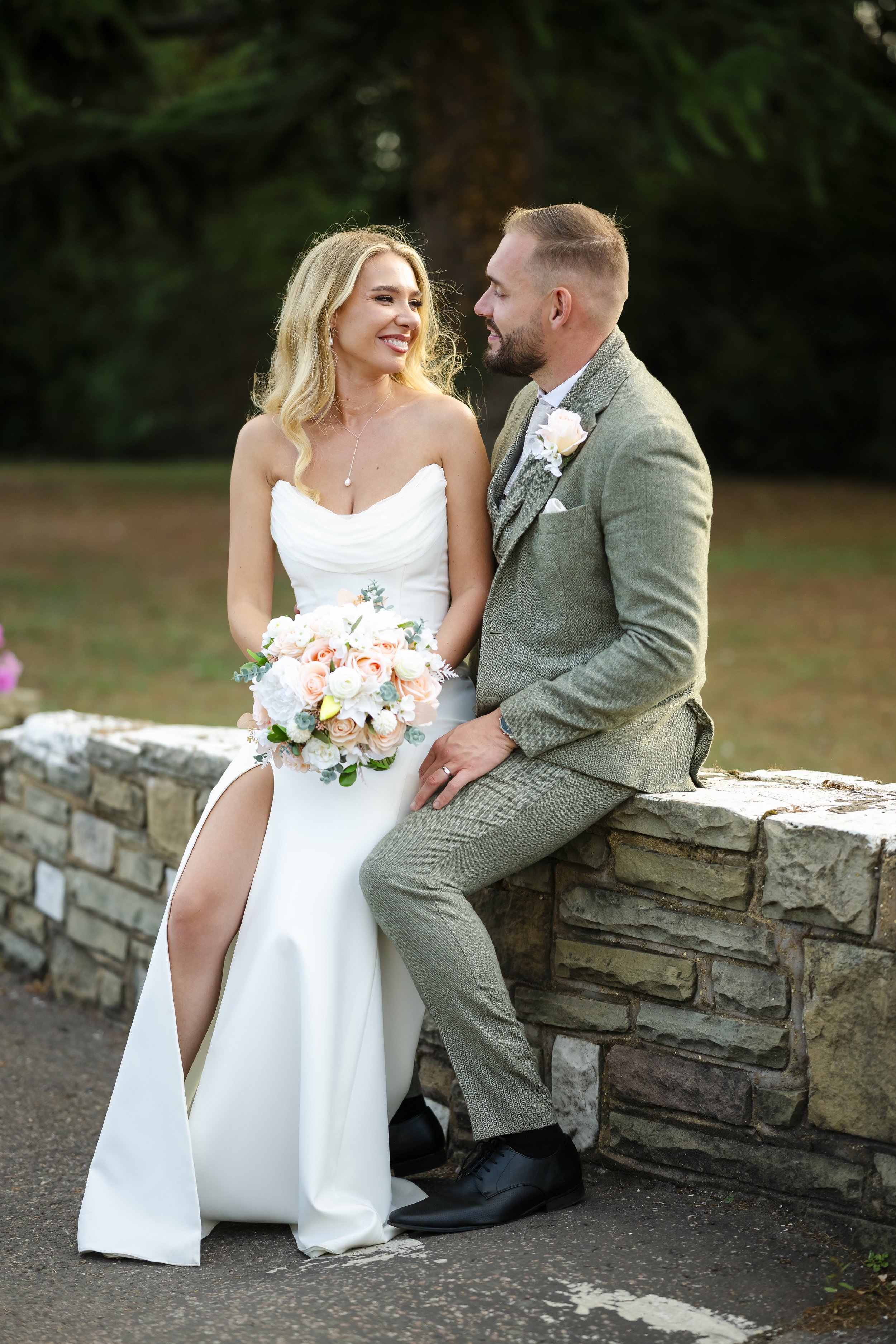 A bride and groom sitting on a stone wall outdoors during their wedding, smiling and looking at each other. The bride is holding a bouquet of pink and white flowers, wearing a strapless white wedding dress with a slit, and the groom is wearing a gray