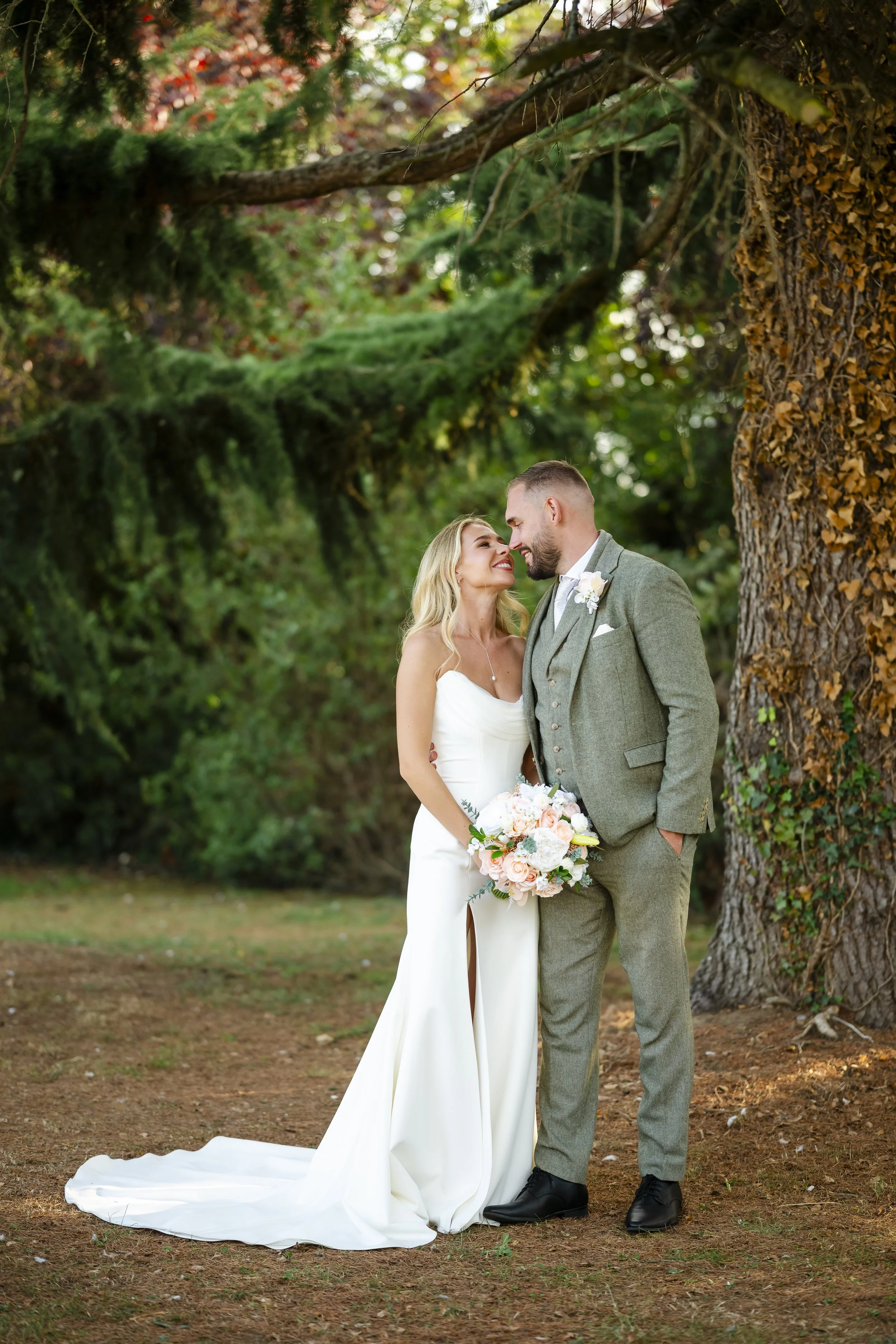 A bride and groom stand close together in a wooded outdoor setting, smiling and gazing into each other's eyes. The bride has long blonde hair, a white wedding dress, and holds a bouquet of pale pink and white flowers. The groom has short hair and a b