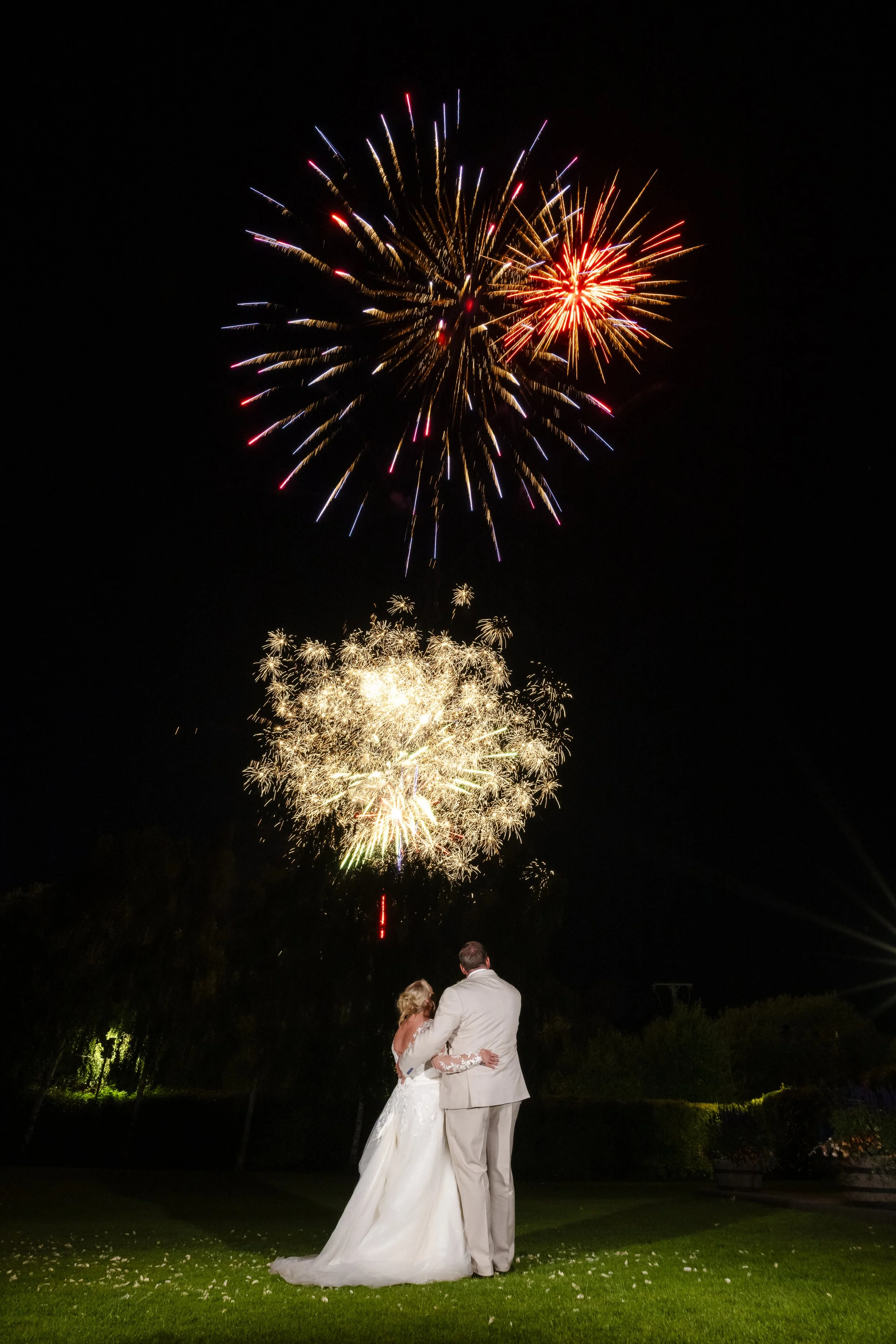A couple dressed in wedding attire looks at fireworks in the night sky during a celebration.