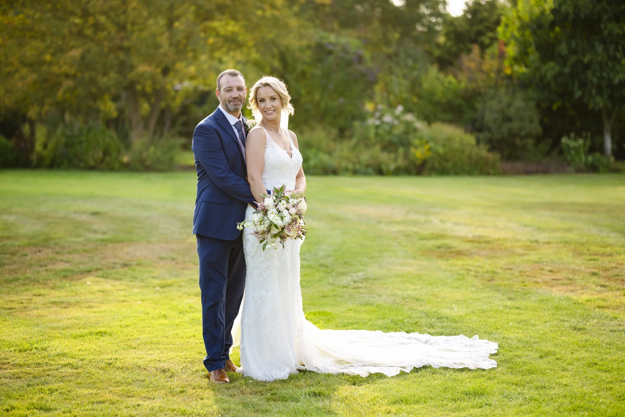 A bride and groom standing together on a grassy field in a park, with trees in the background during sunset, the bride holding a bouquet of flowers, both smiling at the camera.