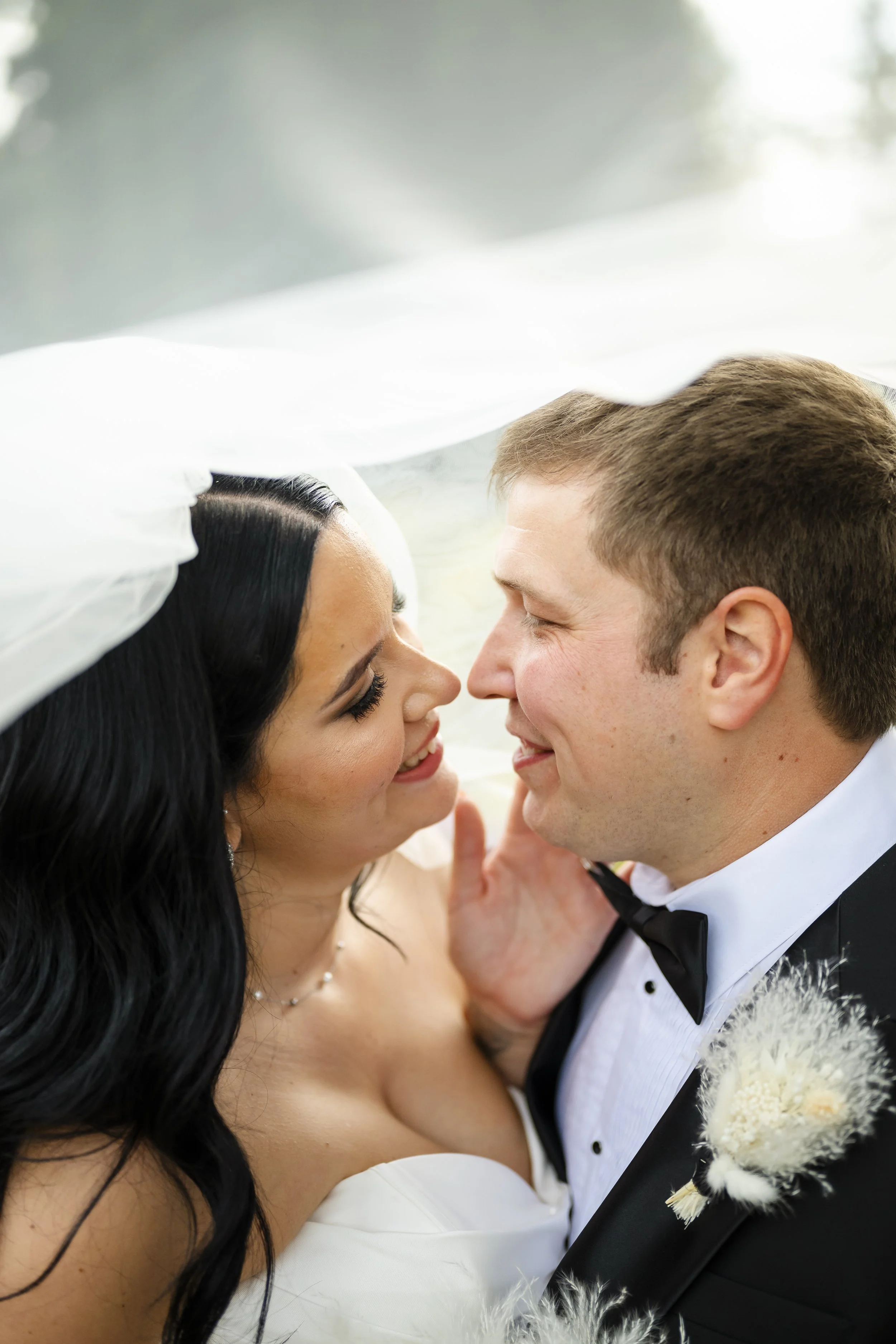 Bride and groom in wedding attire touching noses and gazing at each other under a veil.