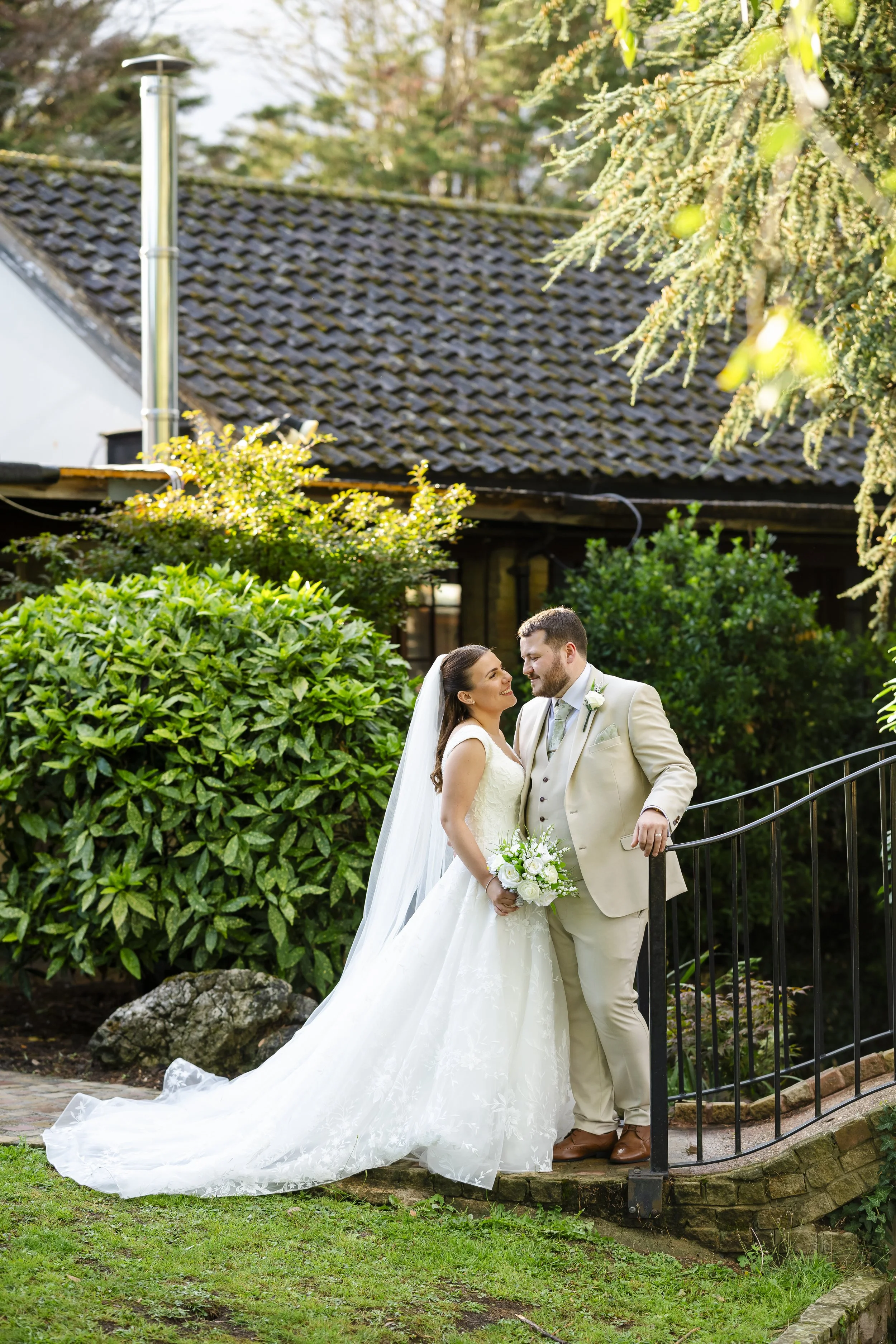 Bride and groom on outdoor staircase smiling at each other, bride in white wedding dress and veil holding bouquet, groom in cream suit with boutonniere, lush green bushes and trees in background.