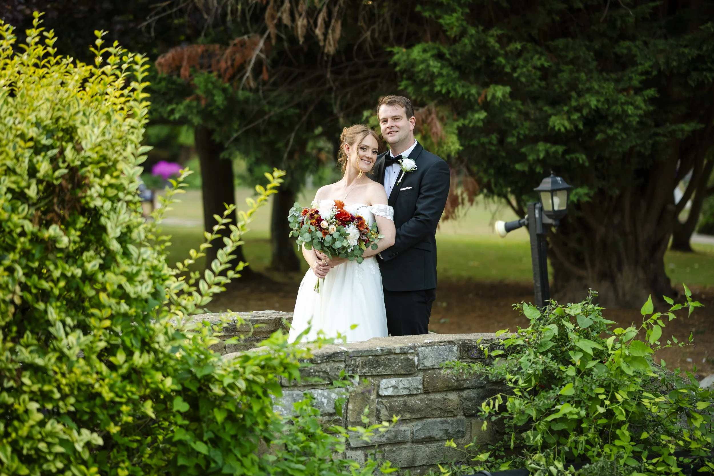 A bride and groom standing outdoors on their wedding day, with lush greenery and trees in the background, the bride holding a bouquet of flowers and the groom dressed in a black tuxedo.
